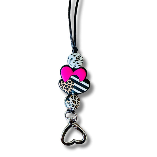 A close up of a simple music lanyard with a leopard print round silicone bead, a pink, leopard print, and black and white striped heart silicone, as well as  a simple white silicone bead with black musical notes on a nylon black string.