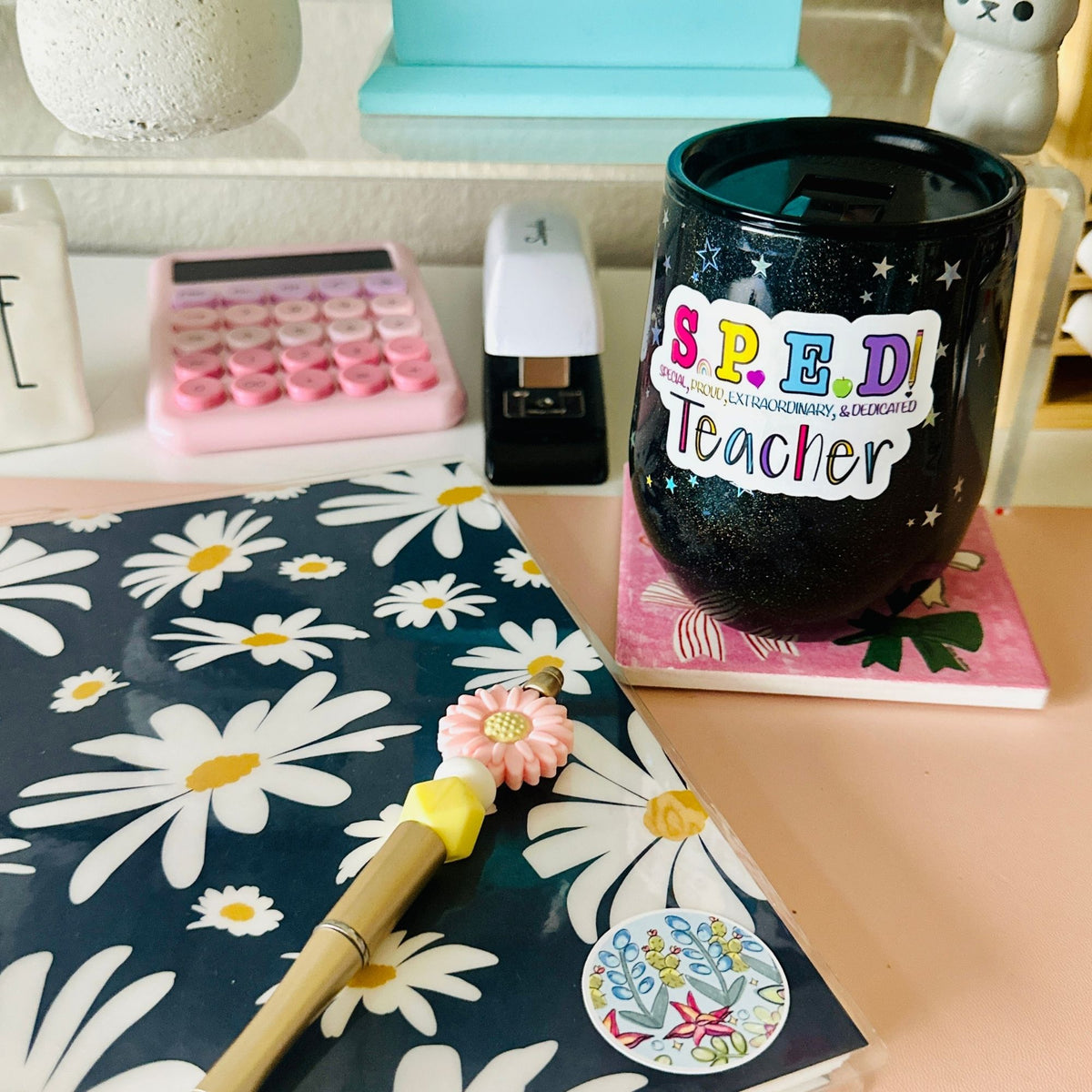 A display of a flowery blue notebook with a flower pen on top next to a starry glitter tumbler with the ‘SPED Teacher’ sticker.