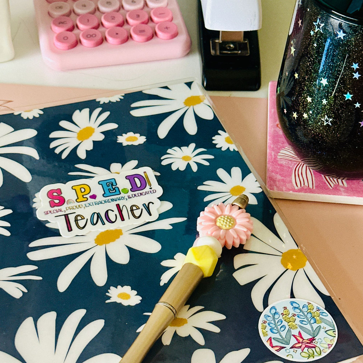A close-up of a floral desk scene featuring a notebook or folder with a navy blue background and large white daisy pattern, along with a ’SPED Teacher’ vinyl sticker in the center.