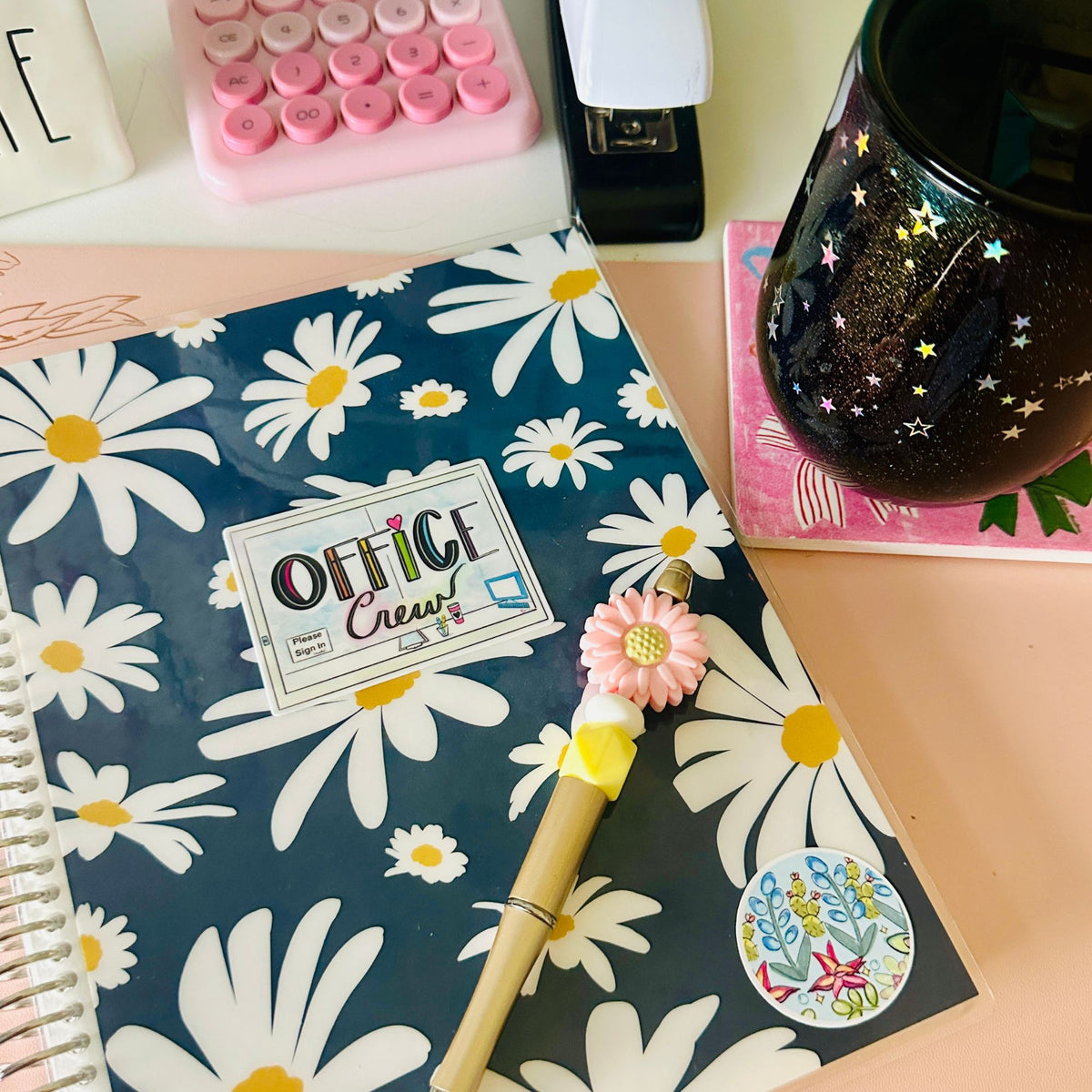 A navy blue planner or notebook with a white daisy floral pattern, featuring a 'Office Crew' sticker on the cover. A pink flower-shaped pen rests on top of the notebook, surrounded by desk accessories including a pink calculator, a black star-patterned mug, and a pink notebook on a peach-colored desk surface.