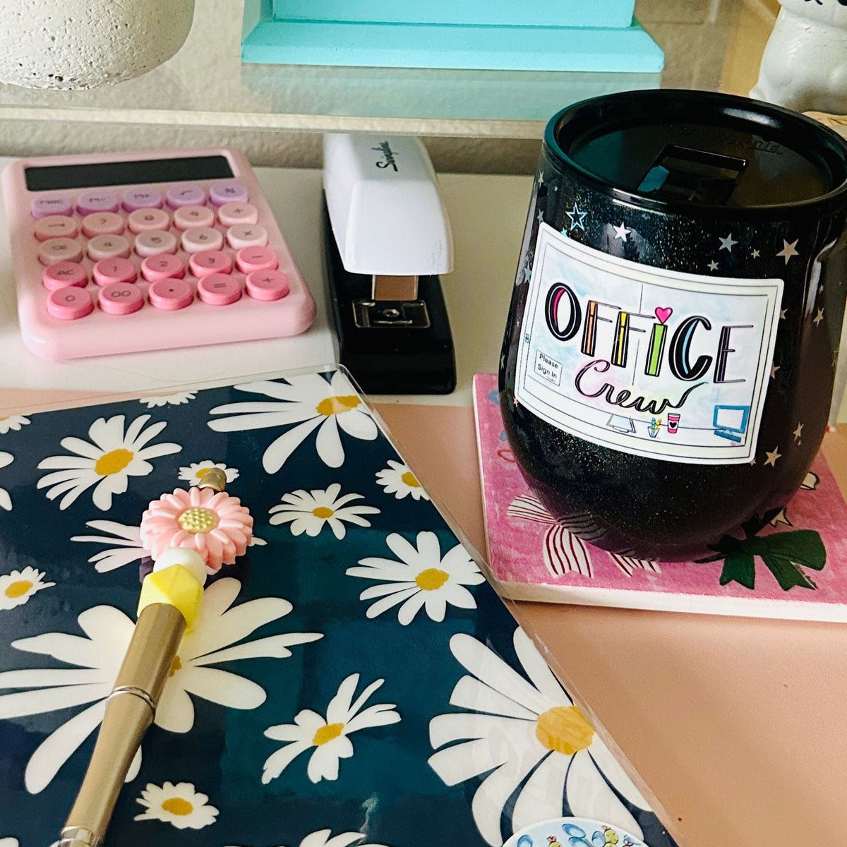 A desk with a floral notebook and pen and next to it is a starry tumbler with the ‘office crew’ vinyl sticker attached.