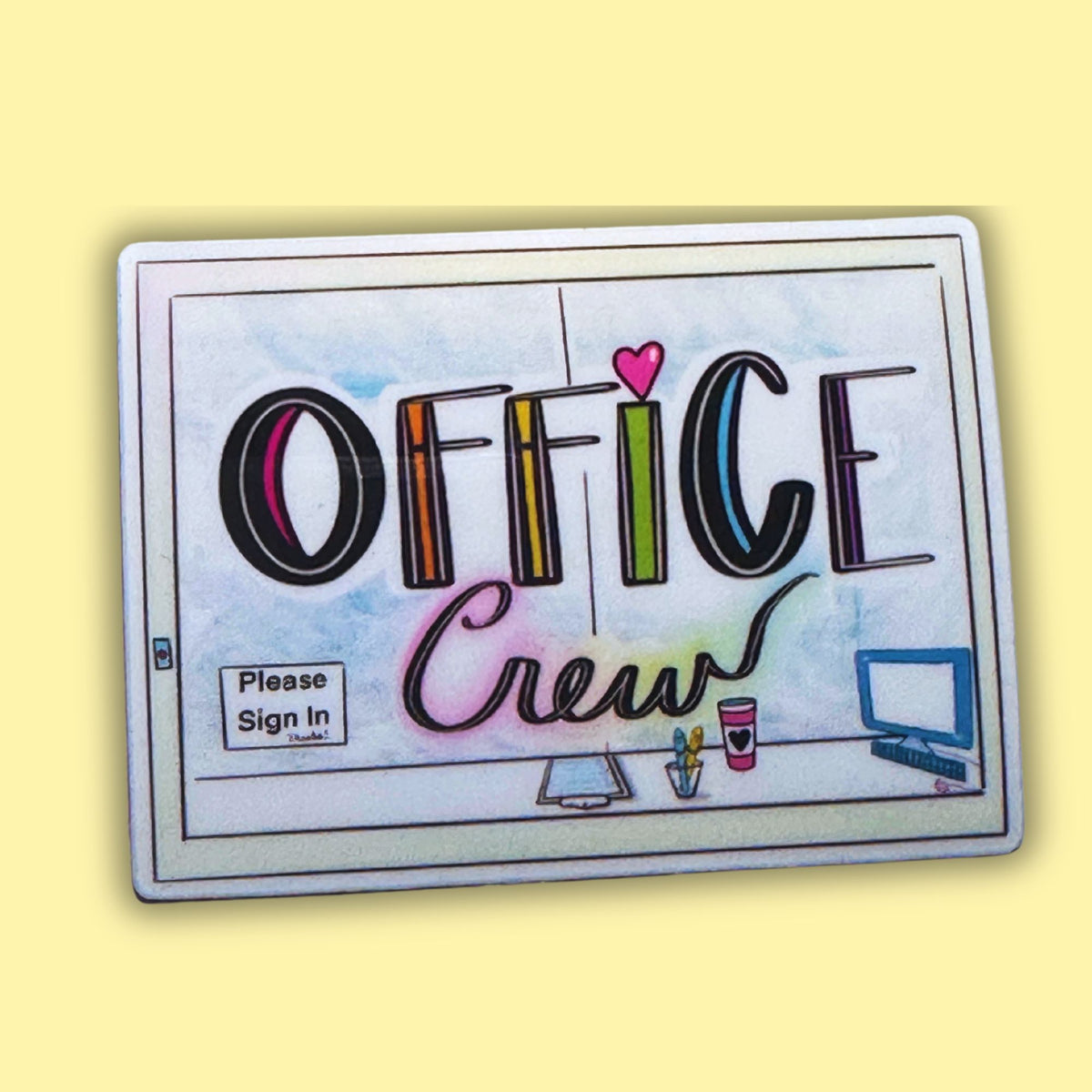 A rectangular printed sign or sticker with rounded corners featuring the words “OFFICE Crew” in stylized multicolor lettering, plus small desk illustrations and a “Please Sign In” box, shown on a pale yellow background.
