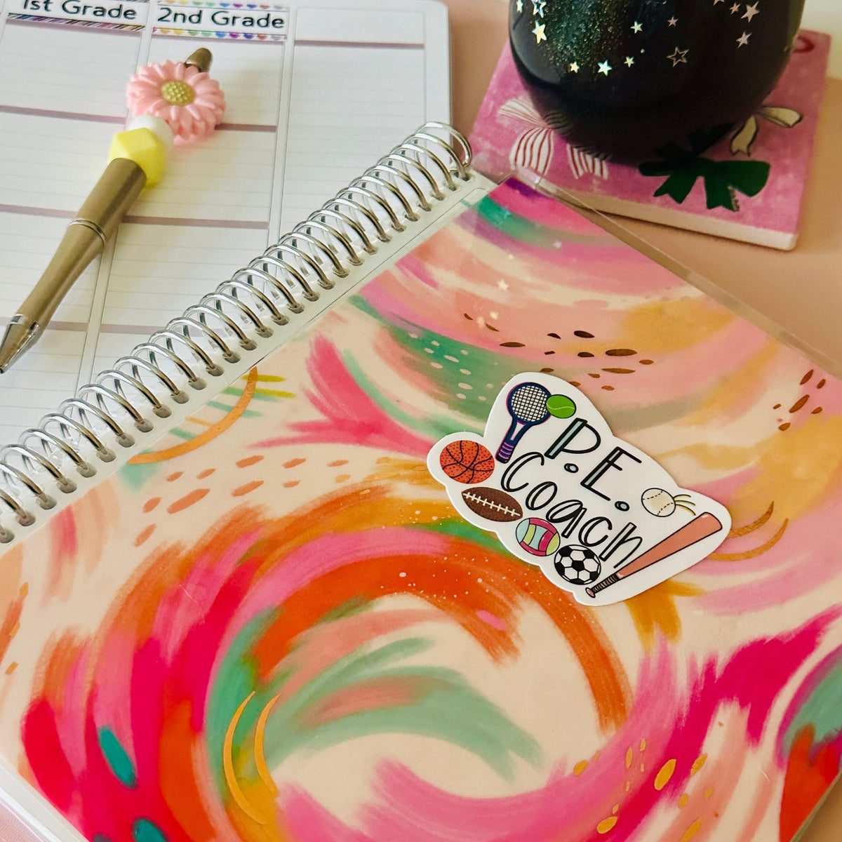 A close-up of a spiral-bound notebook with a colorful abstract paint-splatter cover and a die-cut sticker that reads “P.E. Coach”