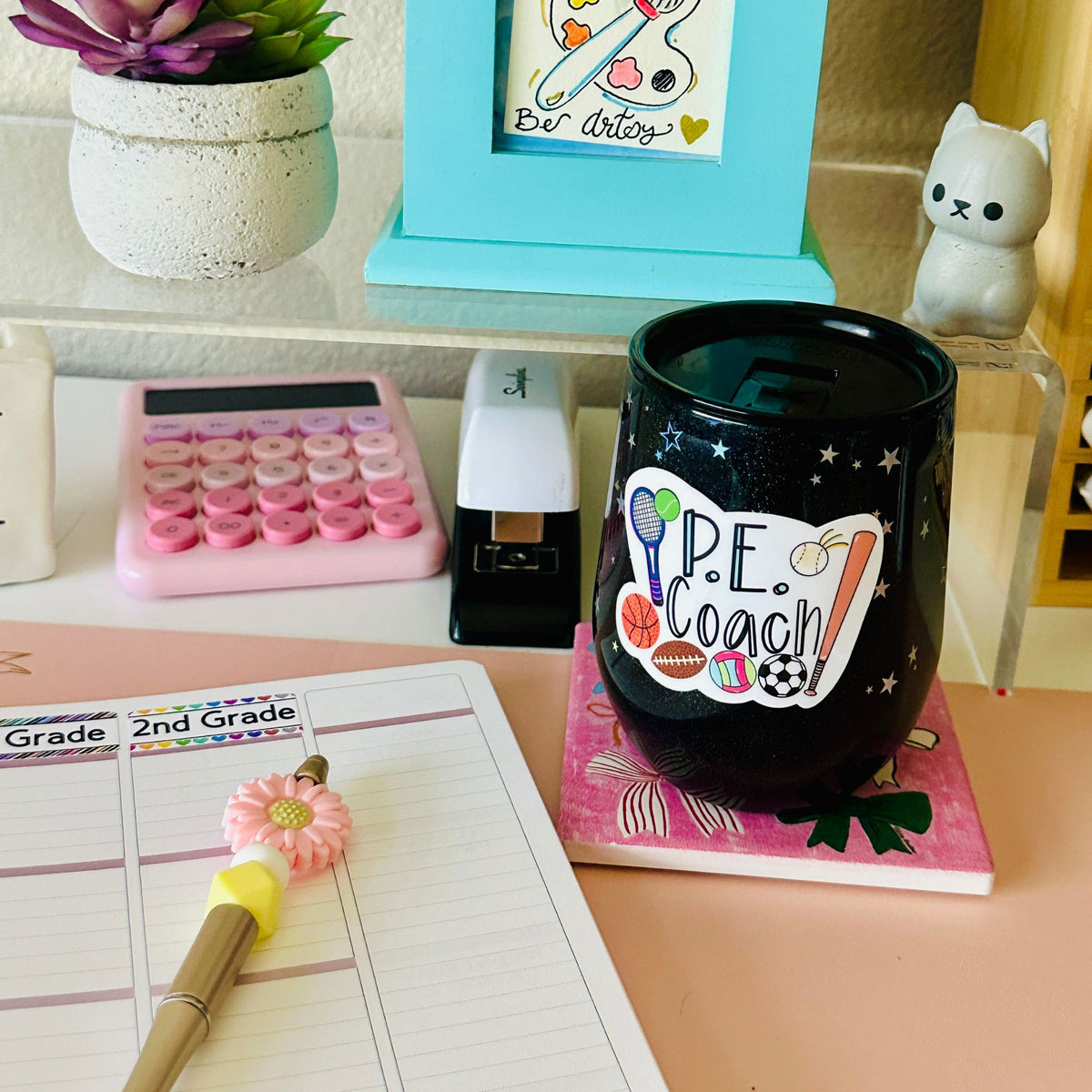 A black shiny tumbler with “P.E. Coach Sticker” resting on a bow coaster with a notepad and pen beside it.