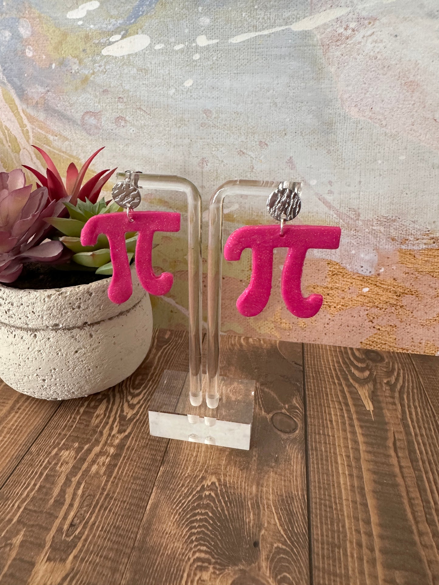 Pink 'Pi' shaped earrings with a stainless steel stud hanging on an acrylic clear earring stand. 