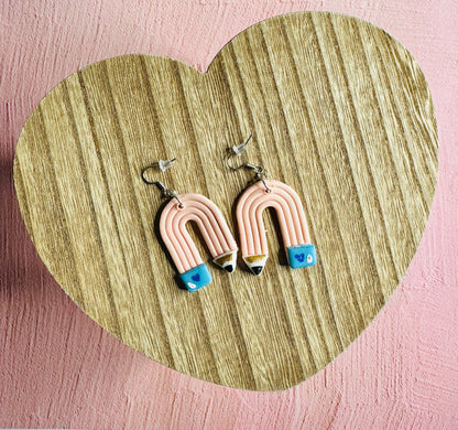 A wide view of the Pink Pencil Teacher Earrings.  The earrings are sitting on a table shaped like a heart.