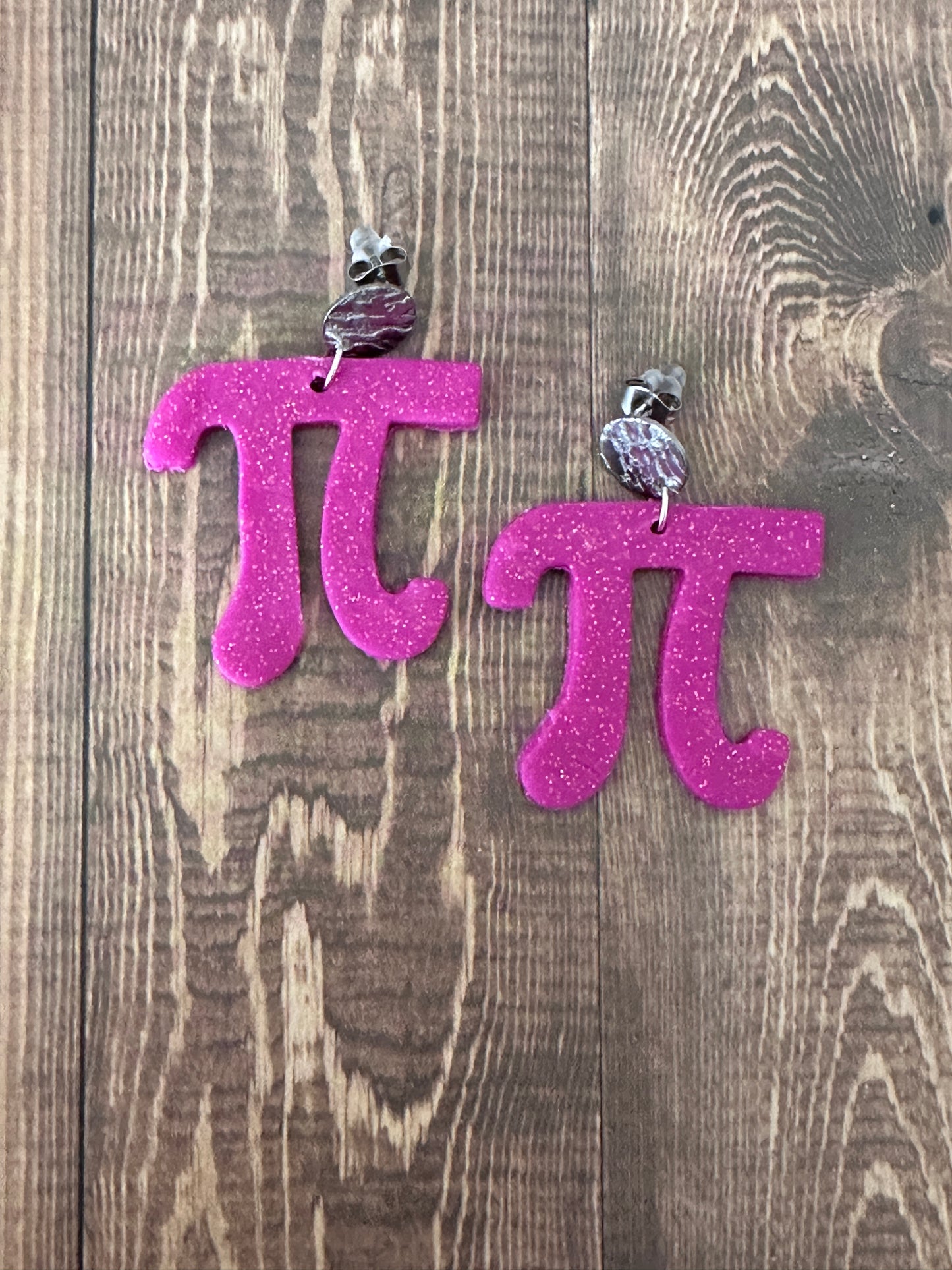 Pink glittery letter 'Pi' earrings on a wooden surface.