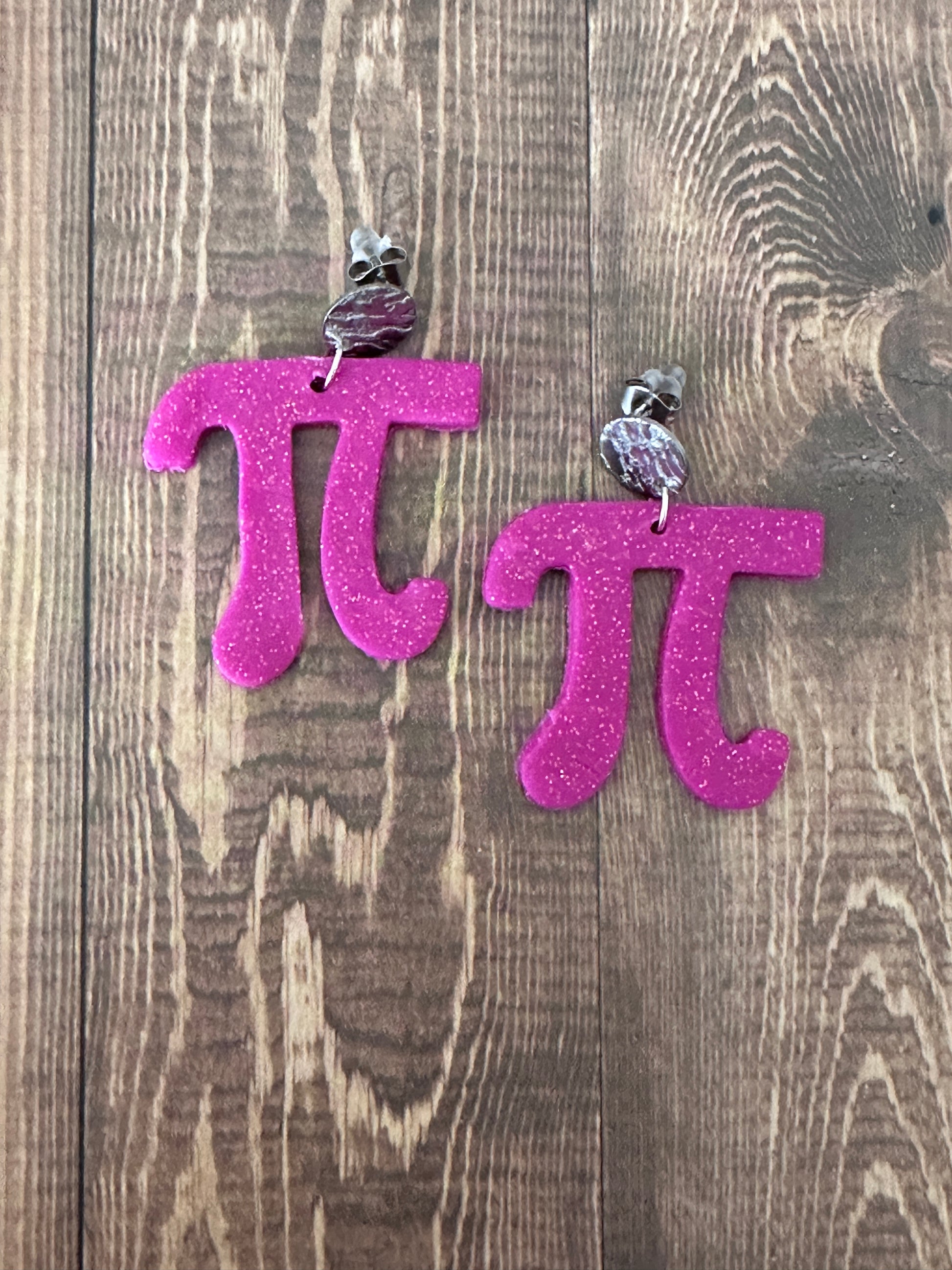 Pink glittery letter 'Pi' earrings on a wooden surface.