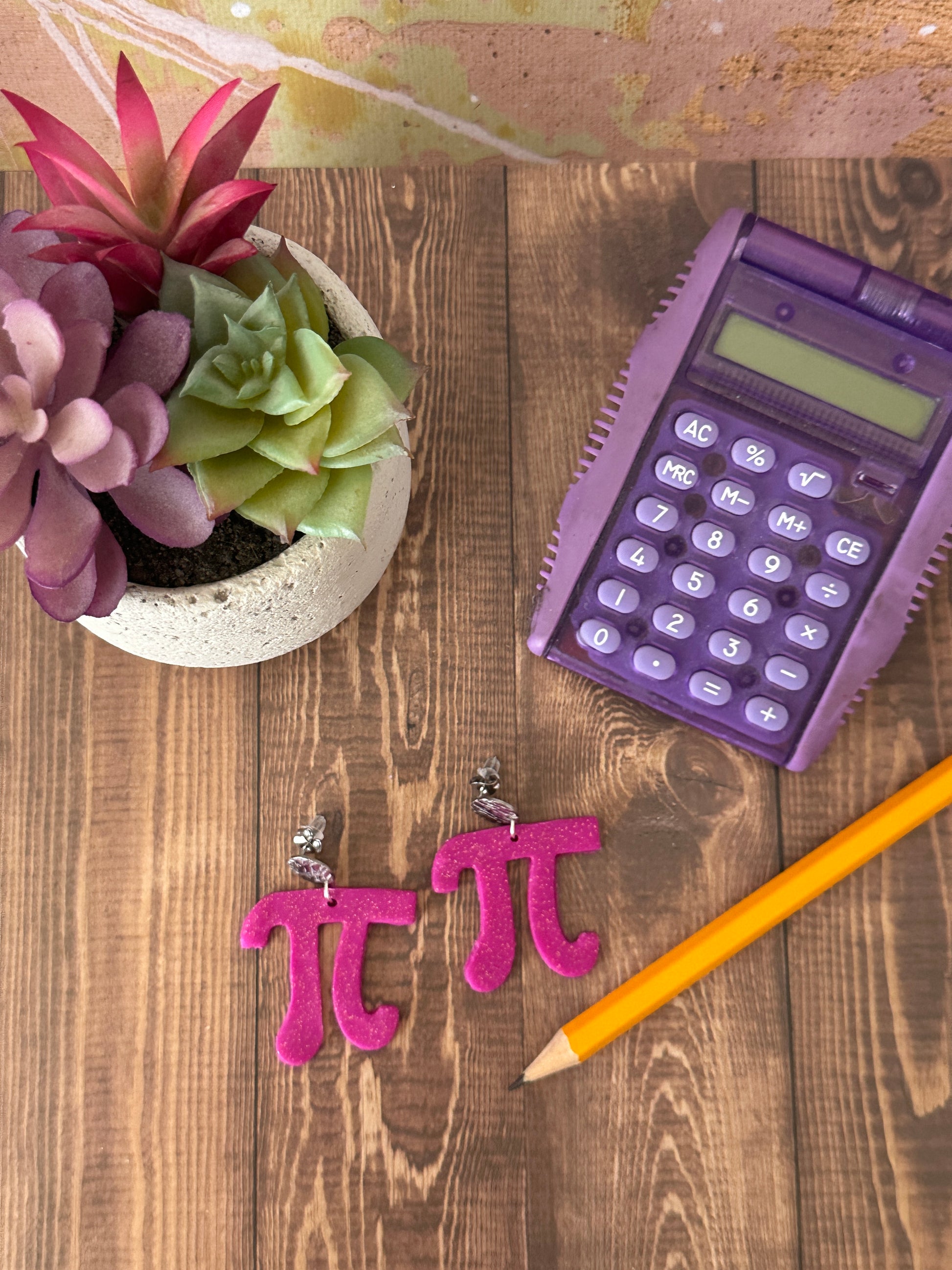 Purple calculator, yellow pencil, pink math pi symbols, small plant on a wooden surface. 