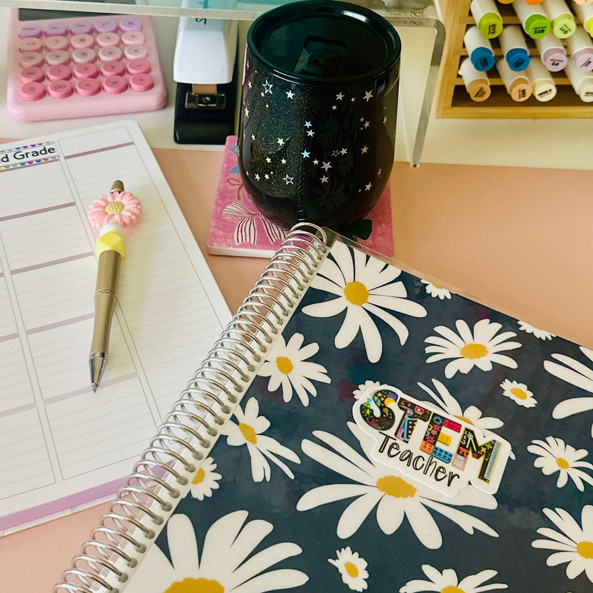 A spiral-bound planner or notebook with a navy blue cover printed with large white daisies and a colorful 'STEM Teacher' sticker, shown on a desk alongside a lined notepad, pen, black cup, and stationery accessories.