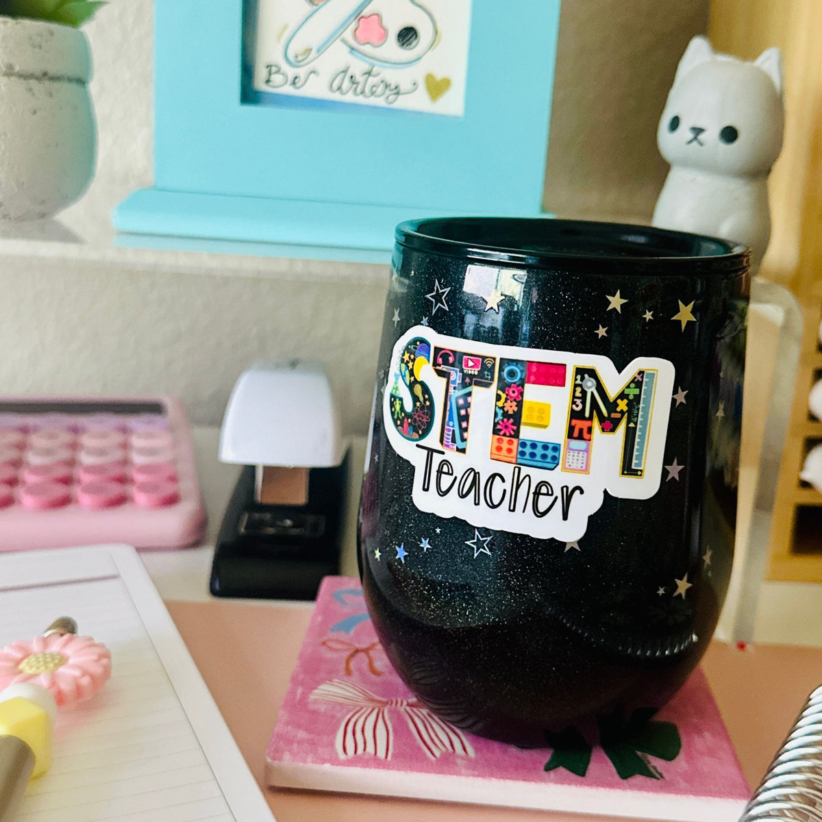 A black glitter stemware tumbler cup with a colorful 'STEM Teacher' sticker, displayed on a desk in a classroom or office setting. 