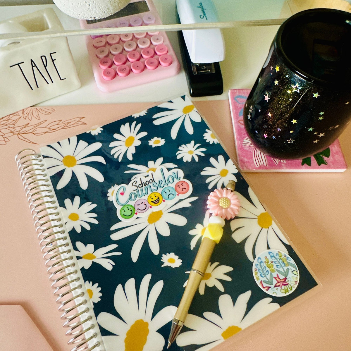 A spiral-bound notebook with a navy blue cover with a white daisy pattern and accessorized with ‘school counselor’ sticker, placed on a pink desk alongside a gold pen, a black glittery mug, and other stationery items.