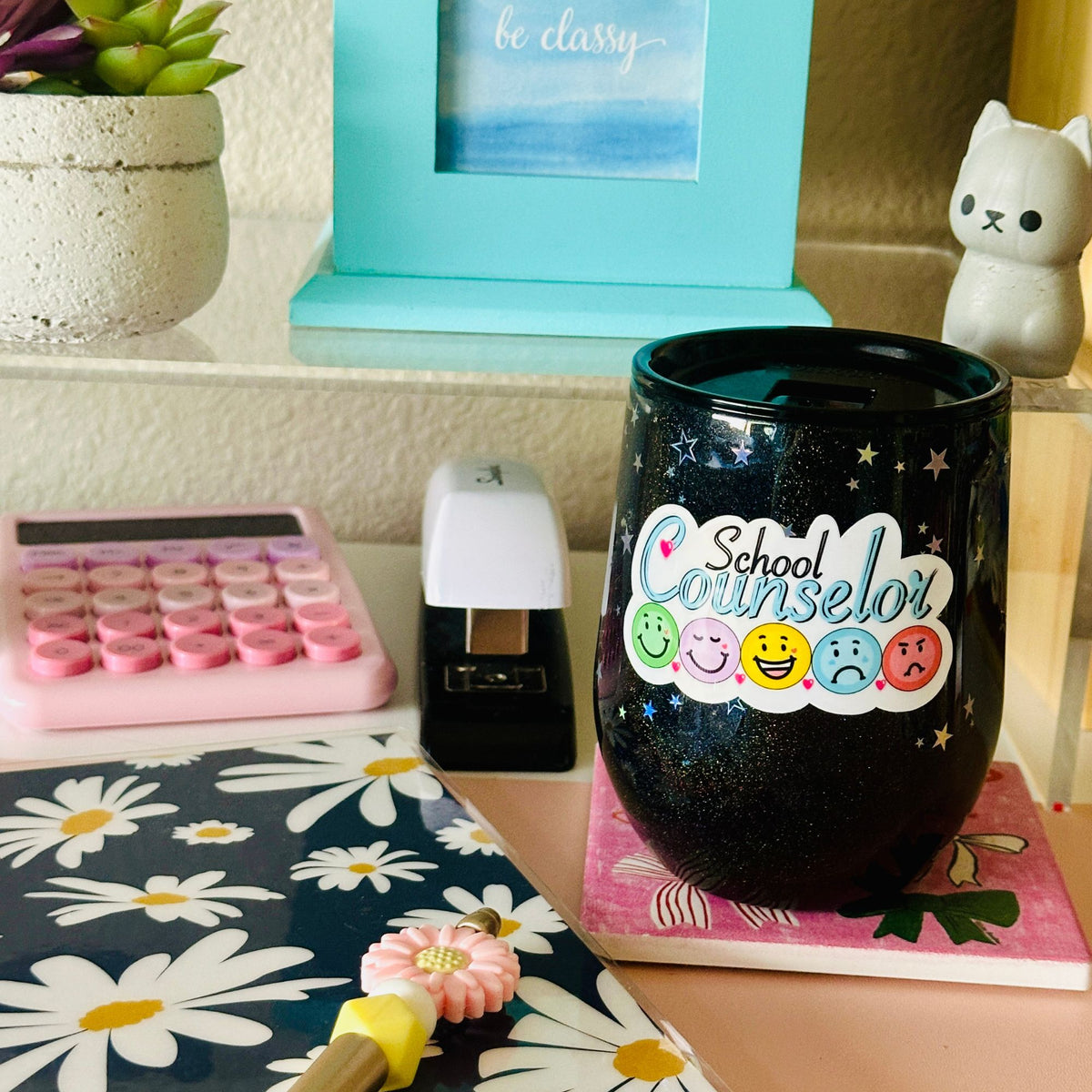 A black glitter tumbler cup with a lid, printed with 'School Counselor' and colorful smiley faces, displayed on a desk with a pink daisy notebook, a pink calculator, and a black stapler.