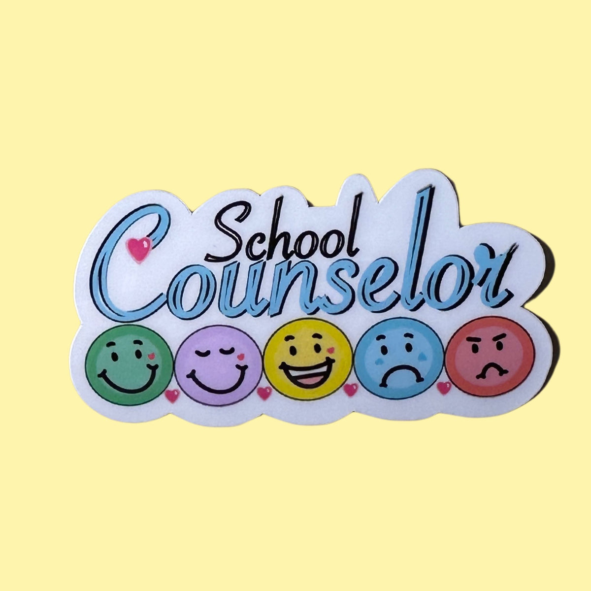 A die-cut sticker or patch with the words 'School Counselor' in stylized blue and black lettering, above five colorful smiley faces representing different emotions, on a pale yellow background.