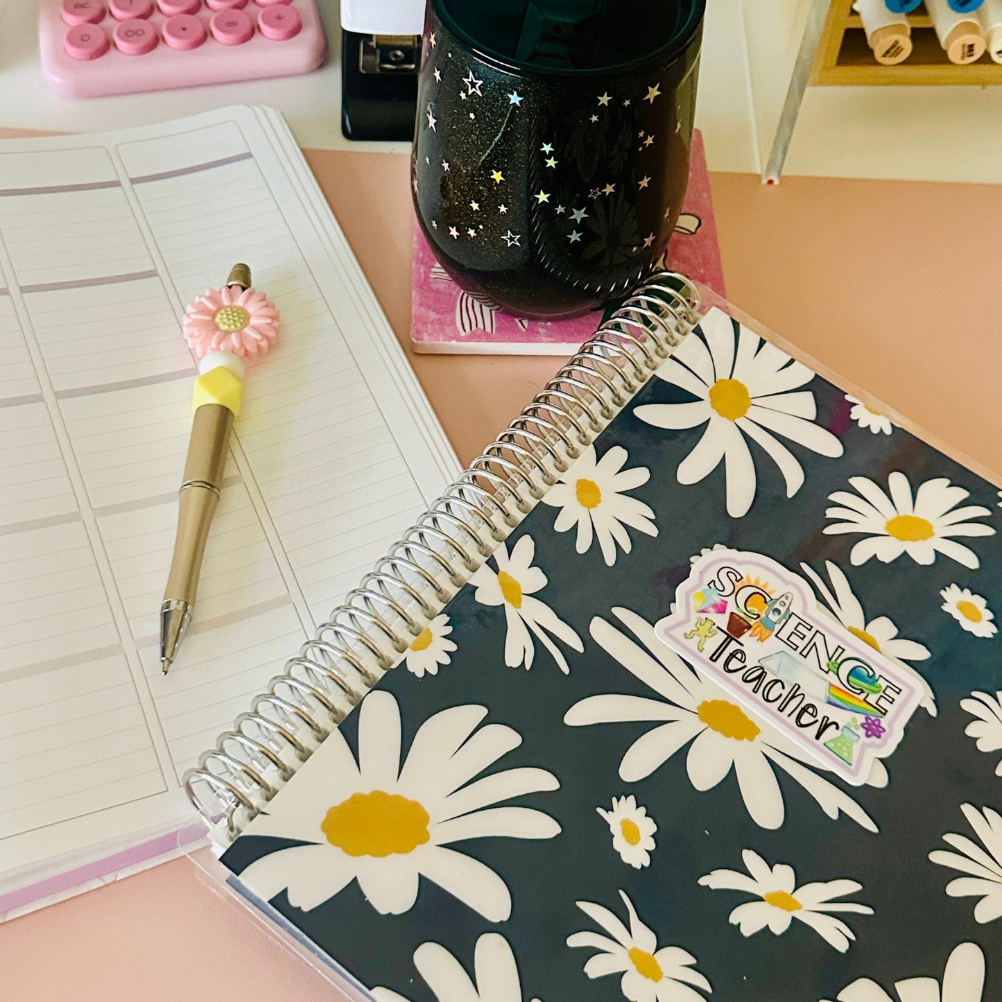 A spiral-bound notebook with a black background and white daisy floral pattern, featuring a 'Science Teacher' sticker on the cover, styled on a desk next to a lined planner page and a pink flower-shaped pen.