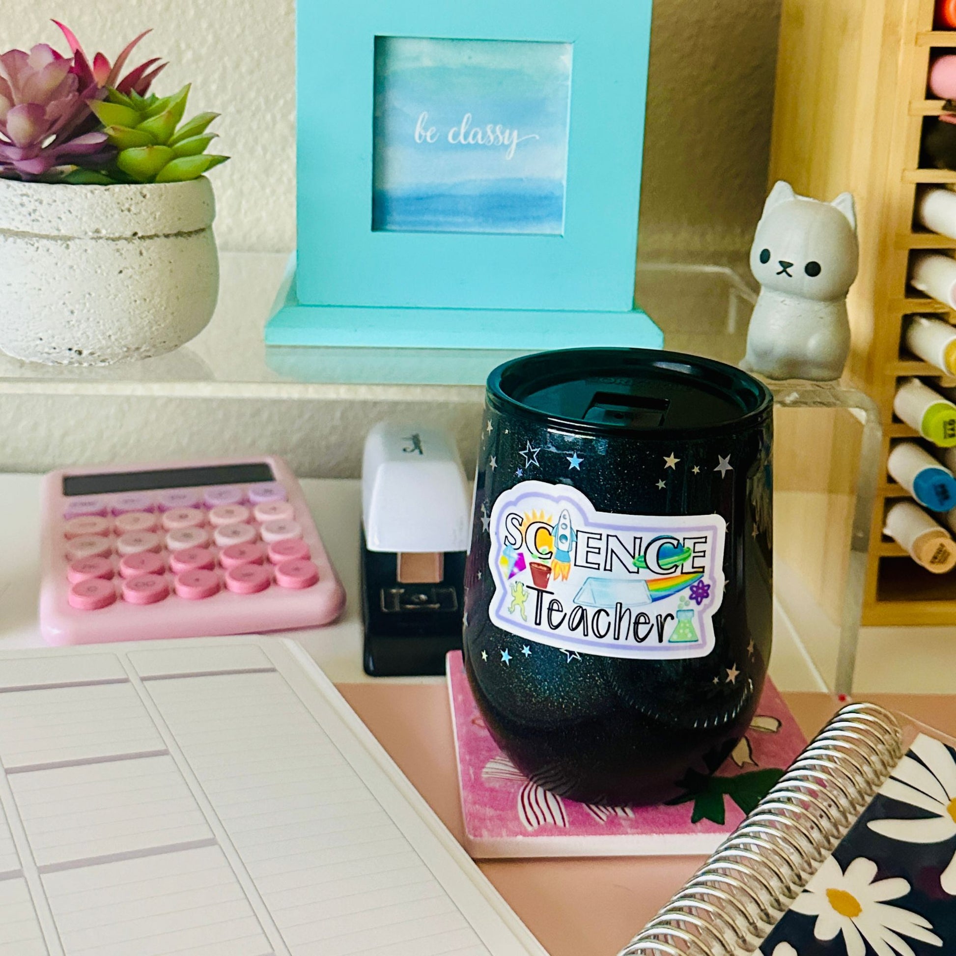 A black glittery stemless wine glass with a 'Science Teacher' sticker, styled on a desk with a pink calculator, notebook, and other school-themed props.