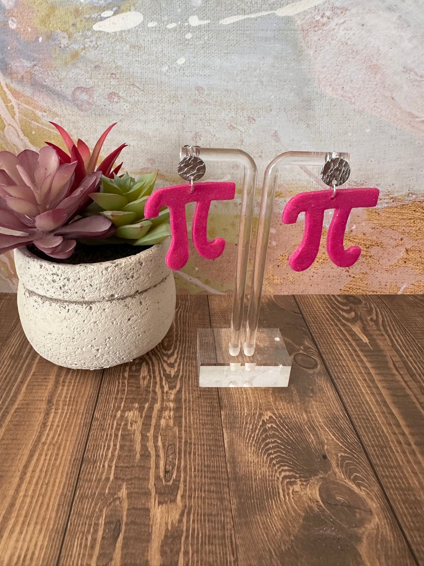 Clear acrylic earring stand holding pink pi shaped earrings resting on wooden surface.