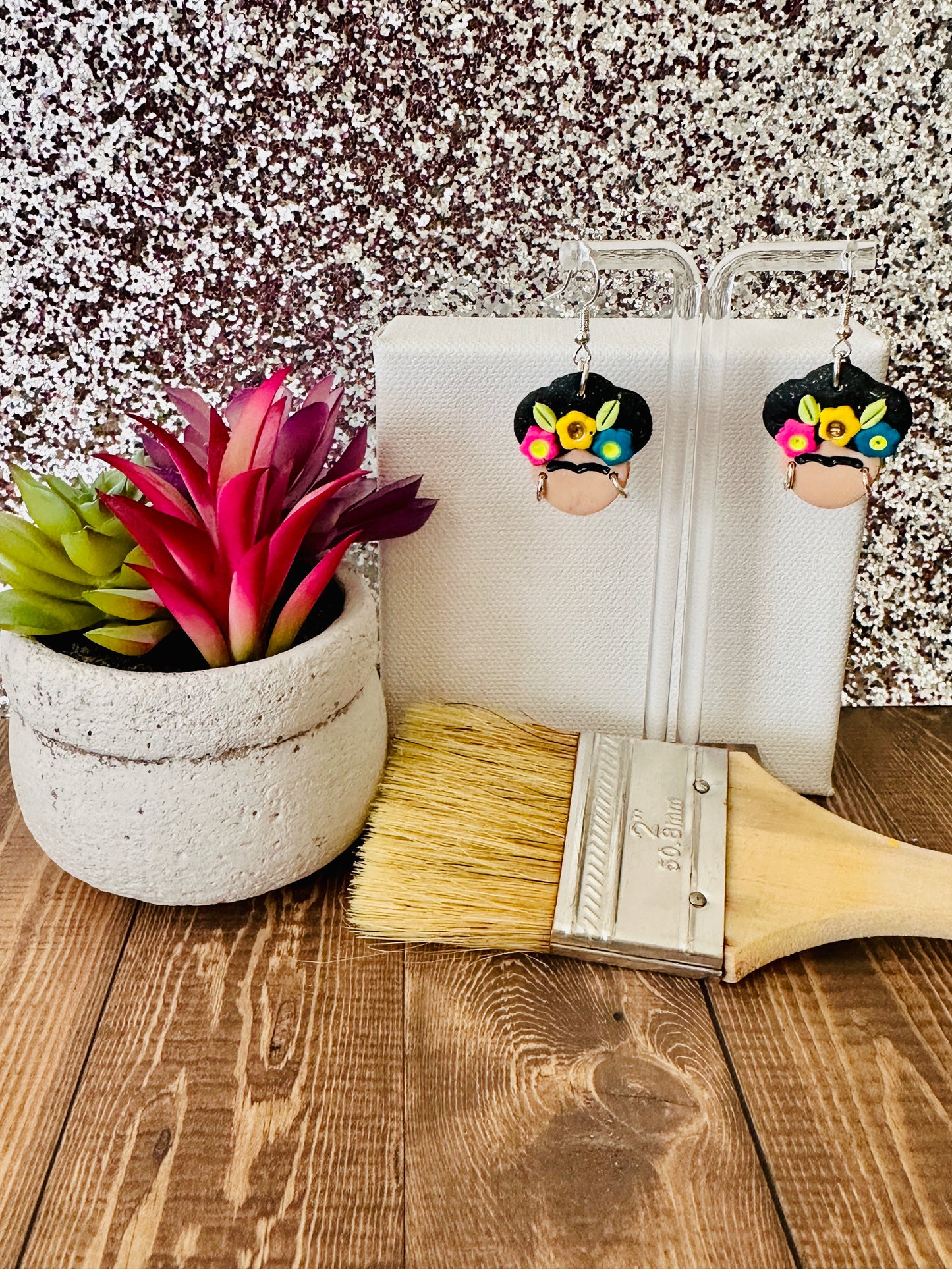 Small Frida Kahlo Bold Flower Crown polymer clay earrings hanging on a clear earring stand.  A paint brush, potted plant, and painter's canvas surround the earrings.  A glittery background and wooden table act as a backdrop.