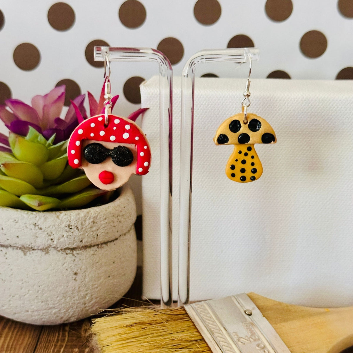 Small variant of the Yayoi Kusama inspired earrings hanging on clear earring stand with polka dot background and paintbrush, showing artistic portrait and mushroom design.