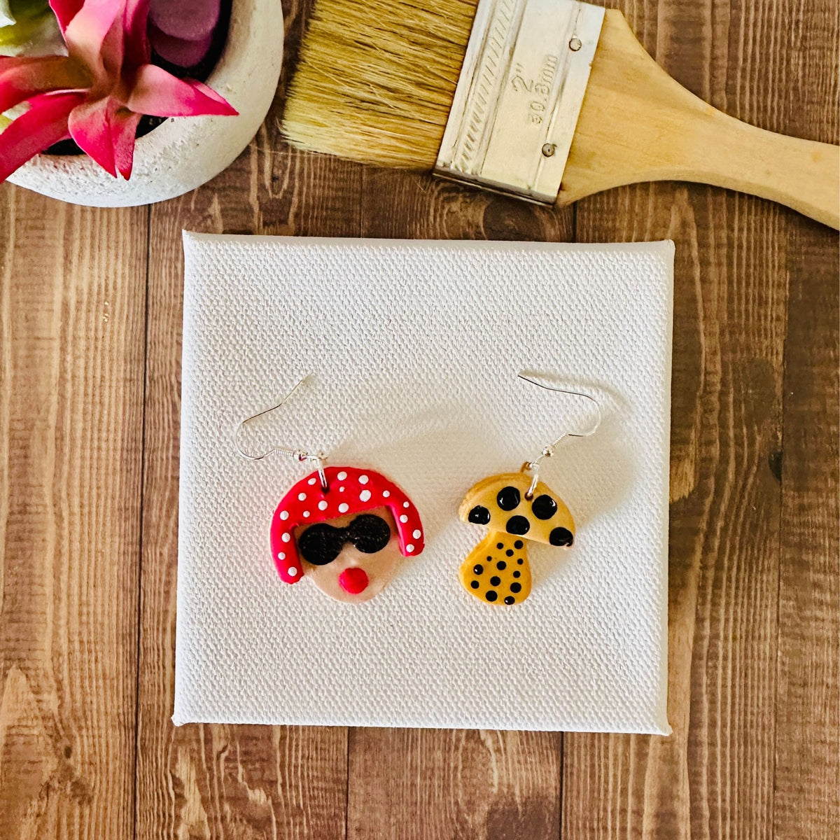 Small Yayoi Kusama polymer clay earrings with portrait featuring red polka dot hair, black sunglasses and yellow mushroom on blank canvas and wooden table.