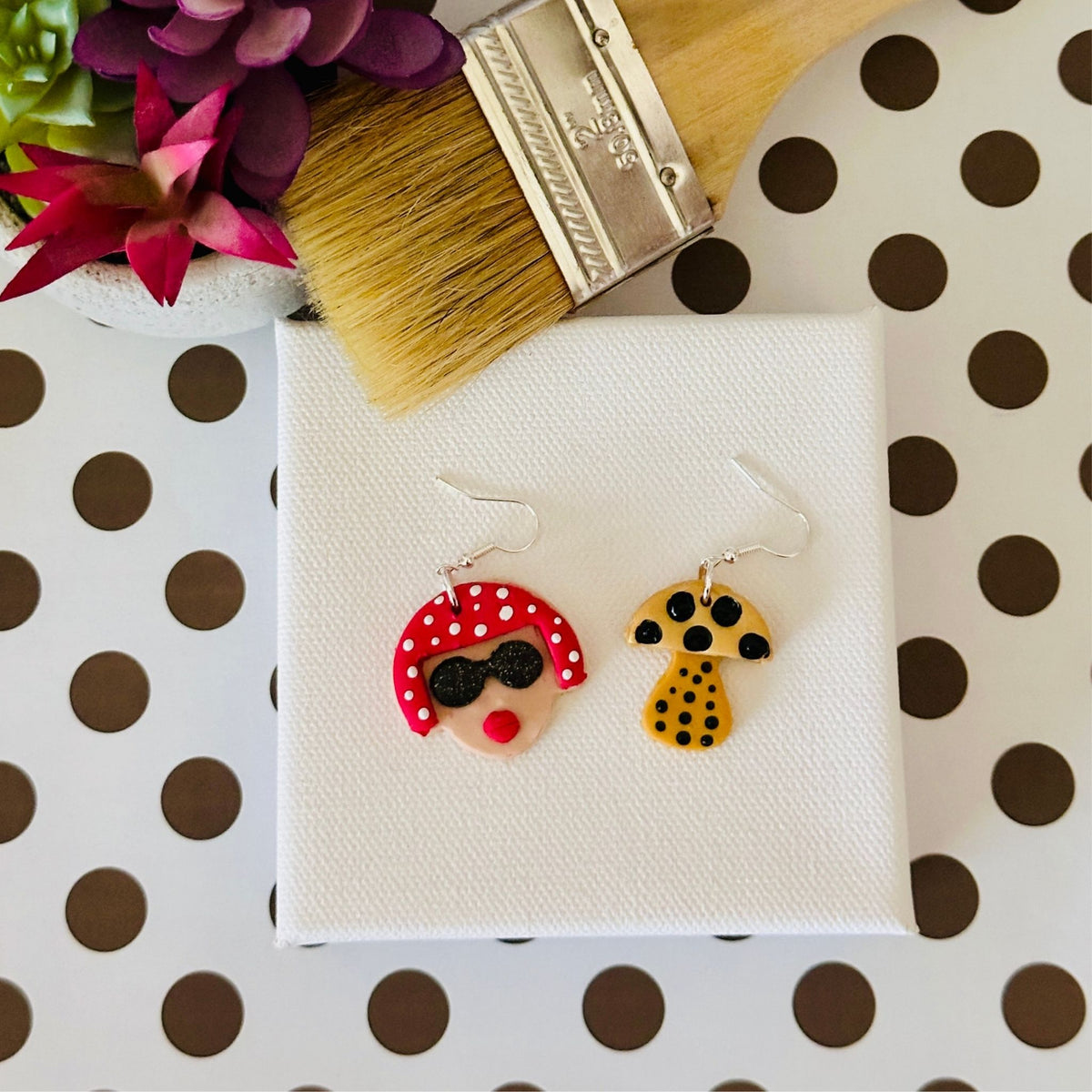 The small variant of the Yayoi Kusama polymer clay earrings displayed on white canvas with paintbrush and potted plant on black and white polka dotted table, perfect for art teachers.