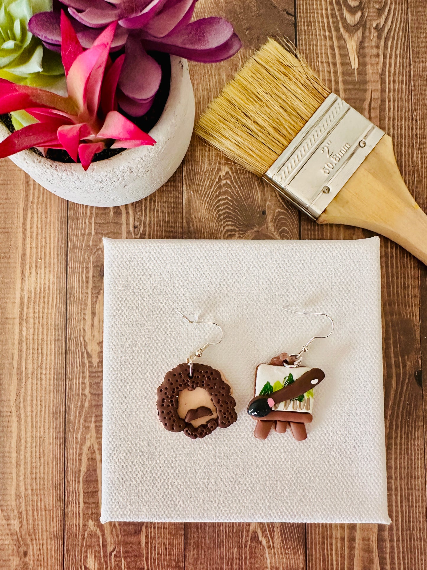 Pair of earrings on a white canvas with a paintbrush and plant in the background on a wooden surface.  This artistic flat lay composition of Bob Ross polymer clay earrings showcases the creative design inspired by the beloved painter and art instructor.