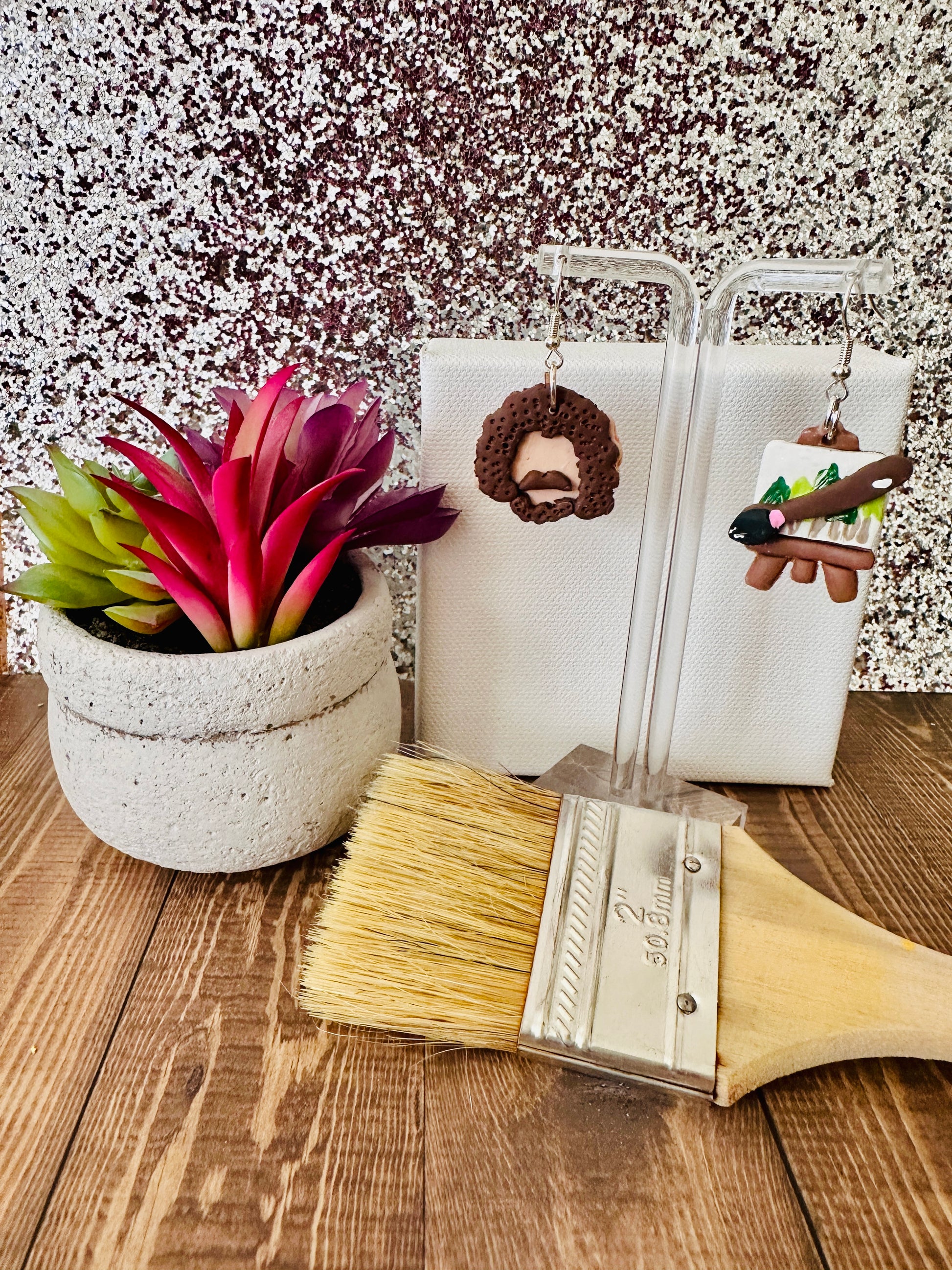 Small Bob Ross earring set displayed by a paint canvas with a paintbrush and a plant on a wooden surface.