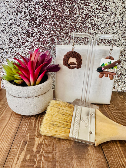 Small Bob Ross earring set displayed by a paint canvas with a paintbrush and a plant on a wooden surface.