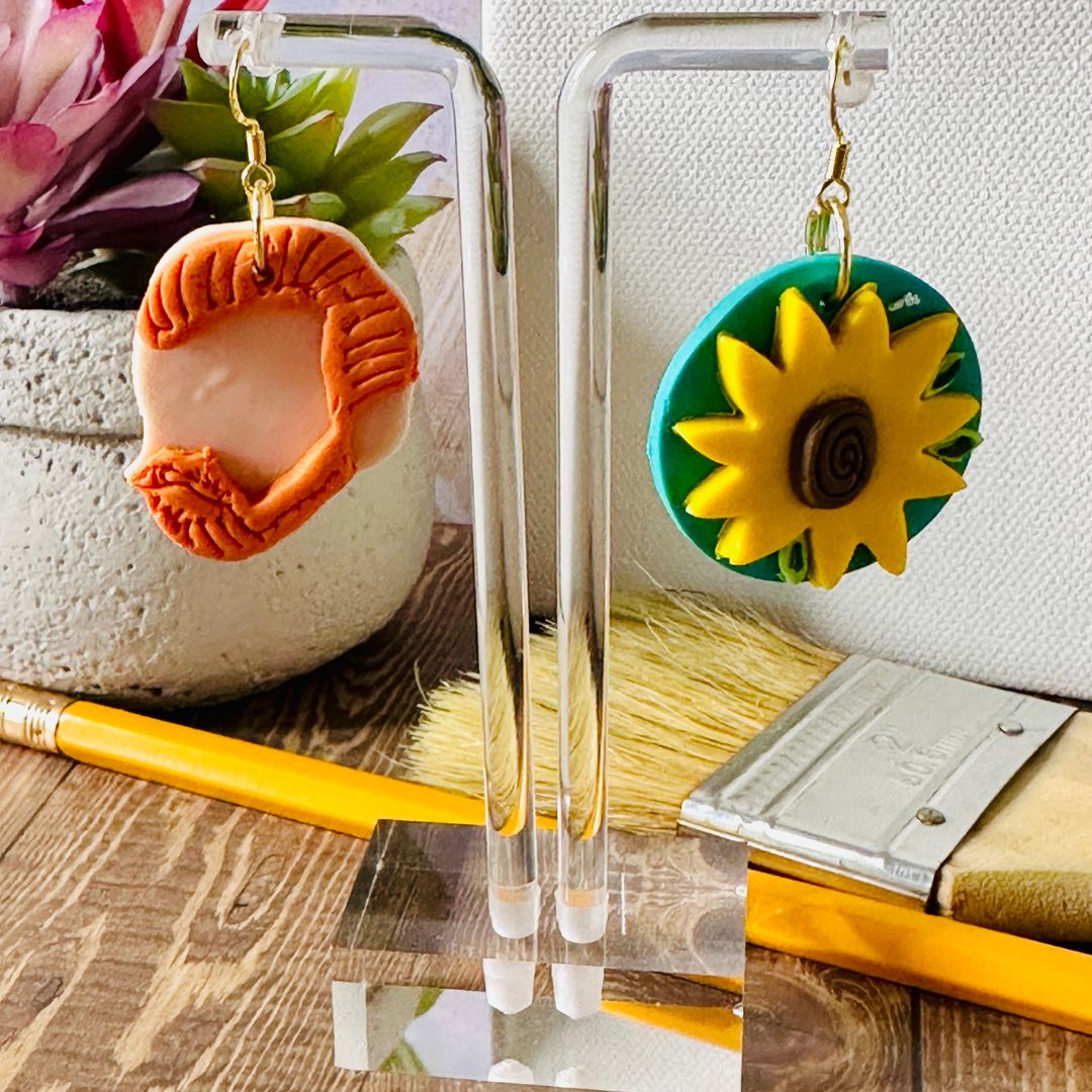 Small Van Gogh art teacher earrings on clear stand with paintbrush, plant on wood table, white canvas wall background.