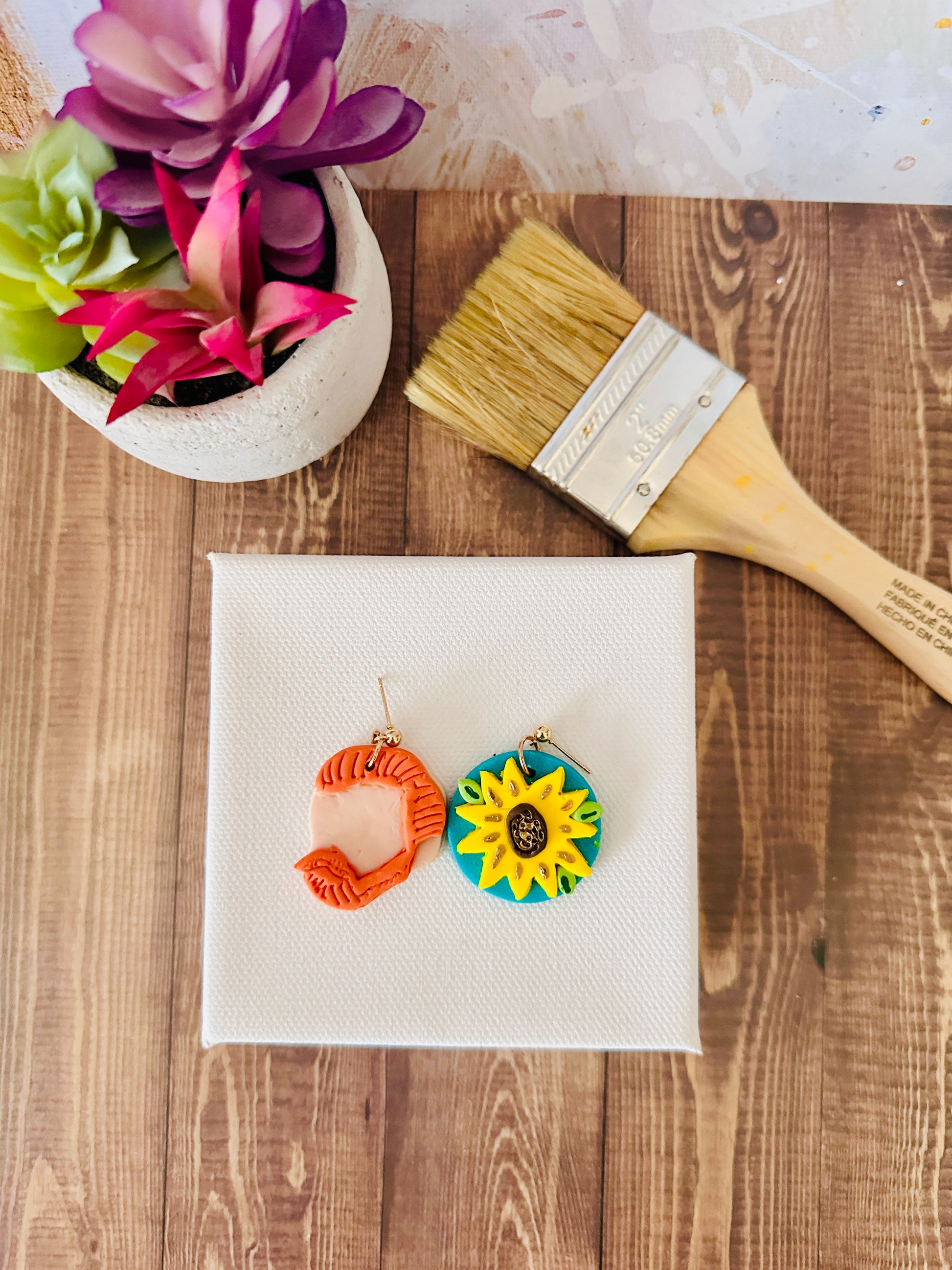 Colorful sunflower earrings on a small white canvas with a paintbrush and plant in the background on a wooden surface.
