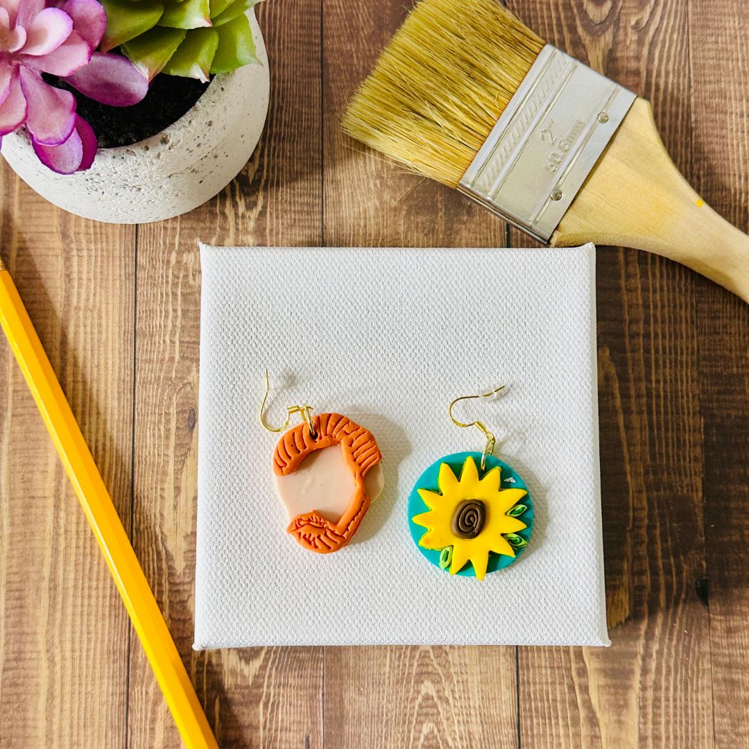 Colorful sunflower earrings on a small white canvas with a paintbrush and plant in the background on a wooden surface.
