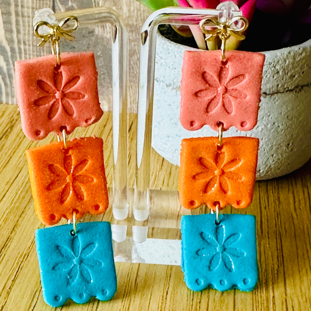 Handmade Spurs pink, teal, and orange papel picado polymer clay earrings with 24k gold bow studs on clear earring stand.
