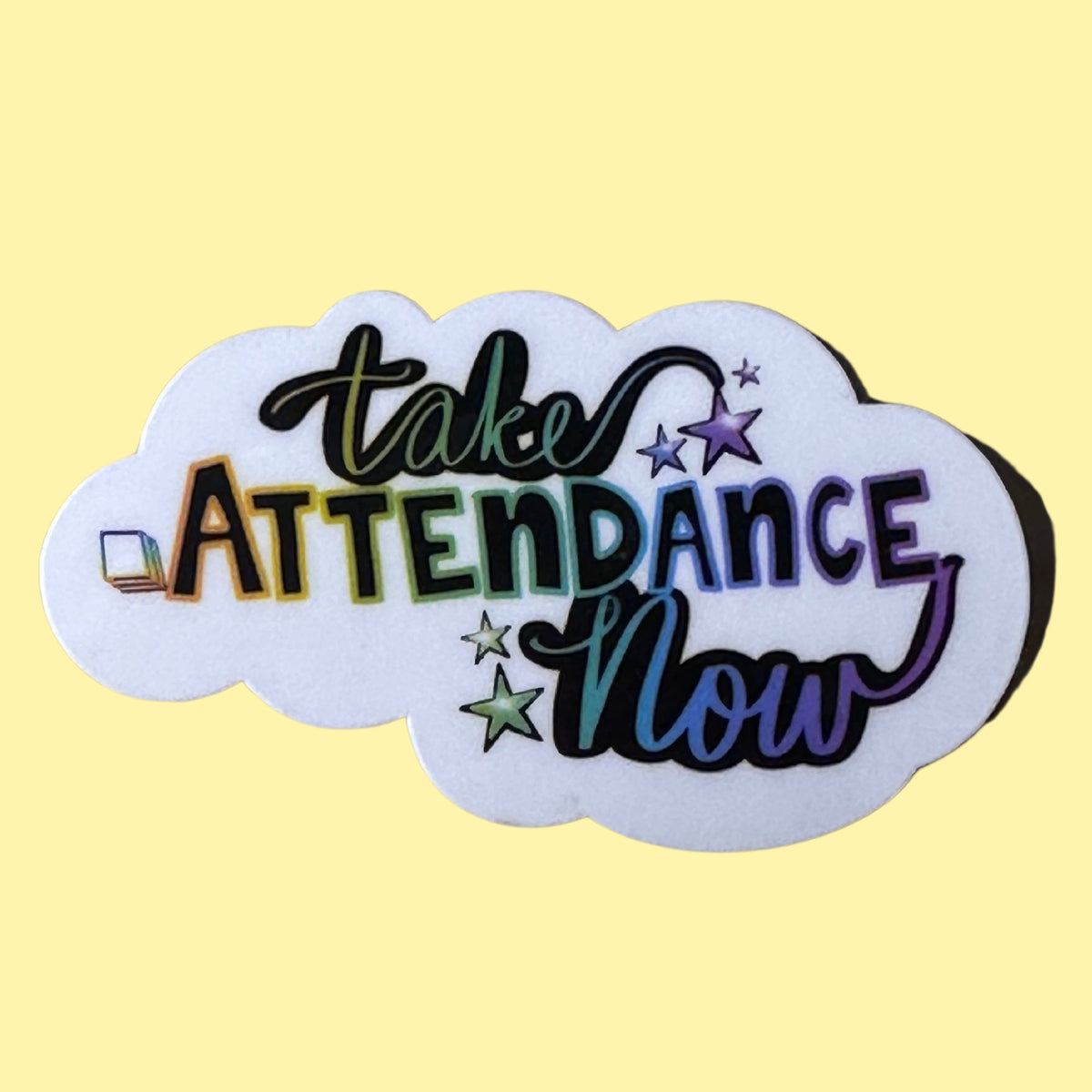 A white cloud-shaped sticker or patch with the phrase 'take ATTENDANCE Now' in multicolored lettering, decorated with small stars and a tiny laptop icon, shown on a pale yellow background.