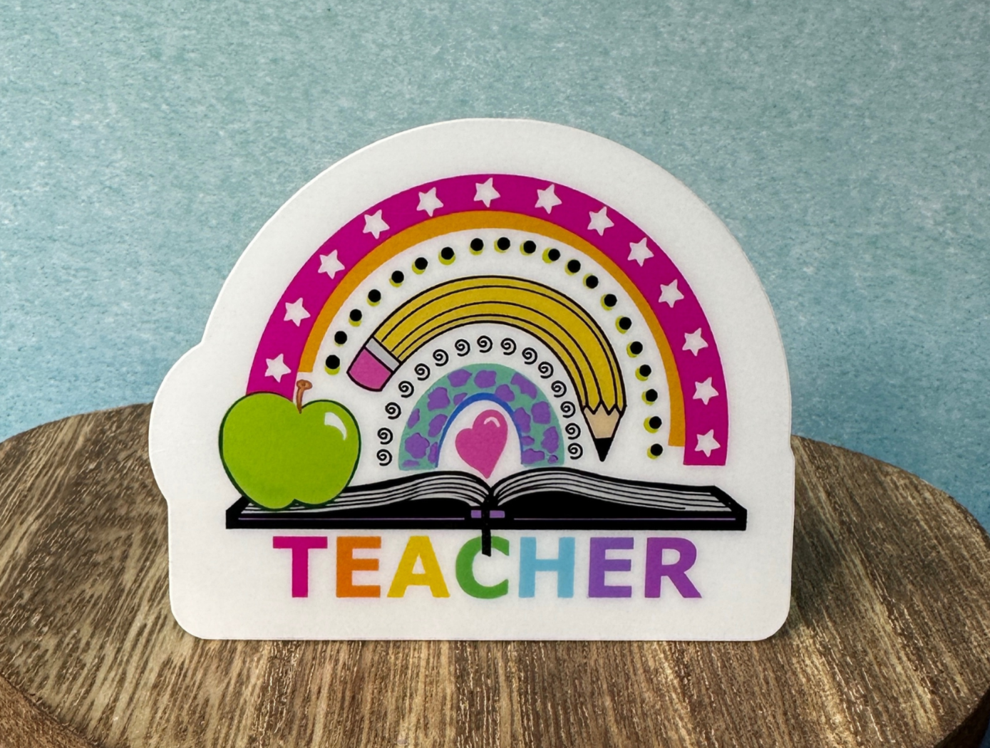 Teacher Rainbow waterproof vinyl sticker displayed on a wooden table in front of a blue wall.