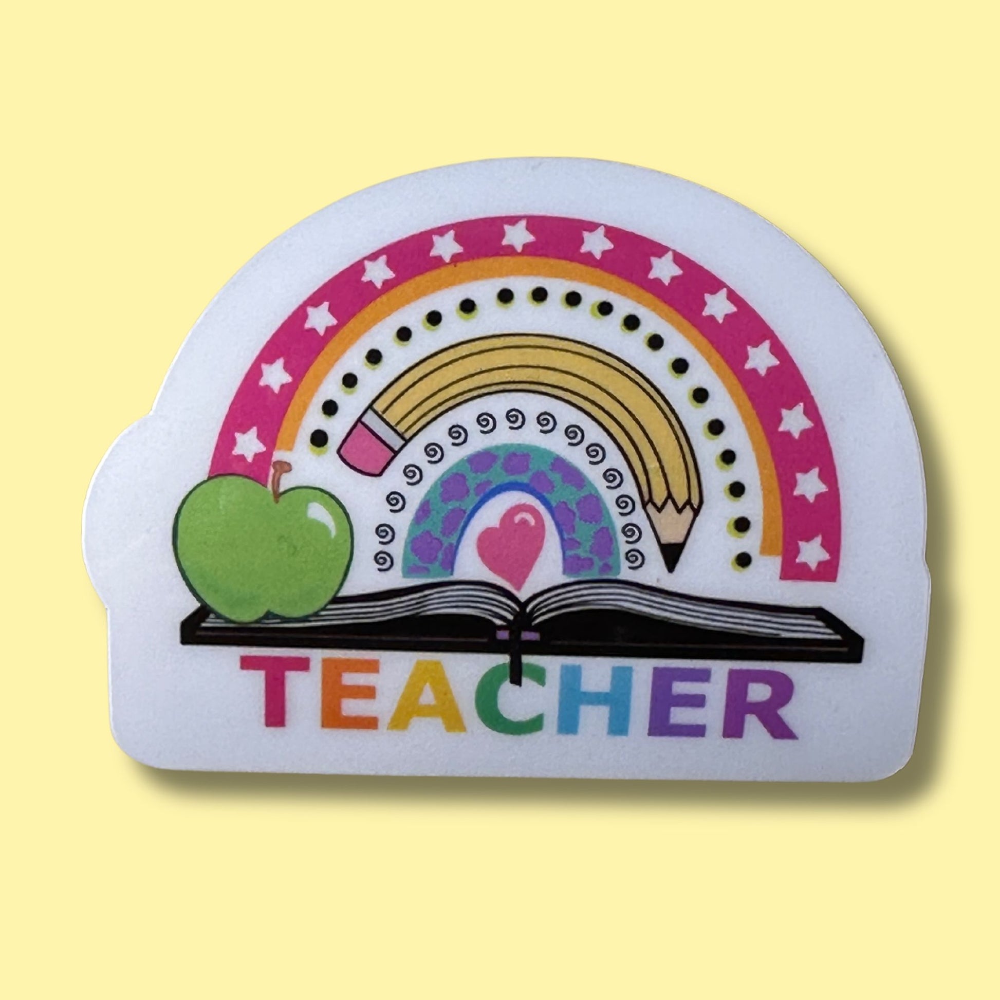 A die-cut white sticker or magnet featuring a colorful illustration of a green apple, open book, rainbow, pencils, stars, and the word 'TEACHER' in rainbow letters, shown on a pale yellow background.