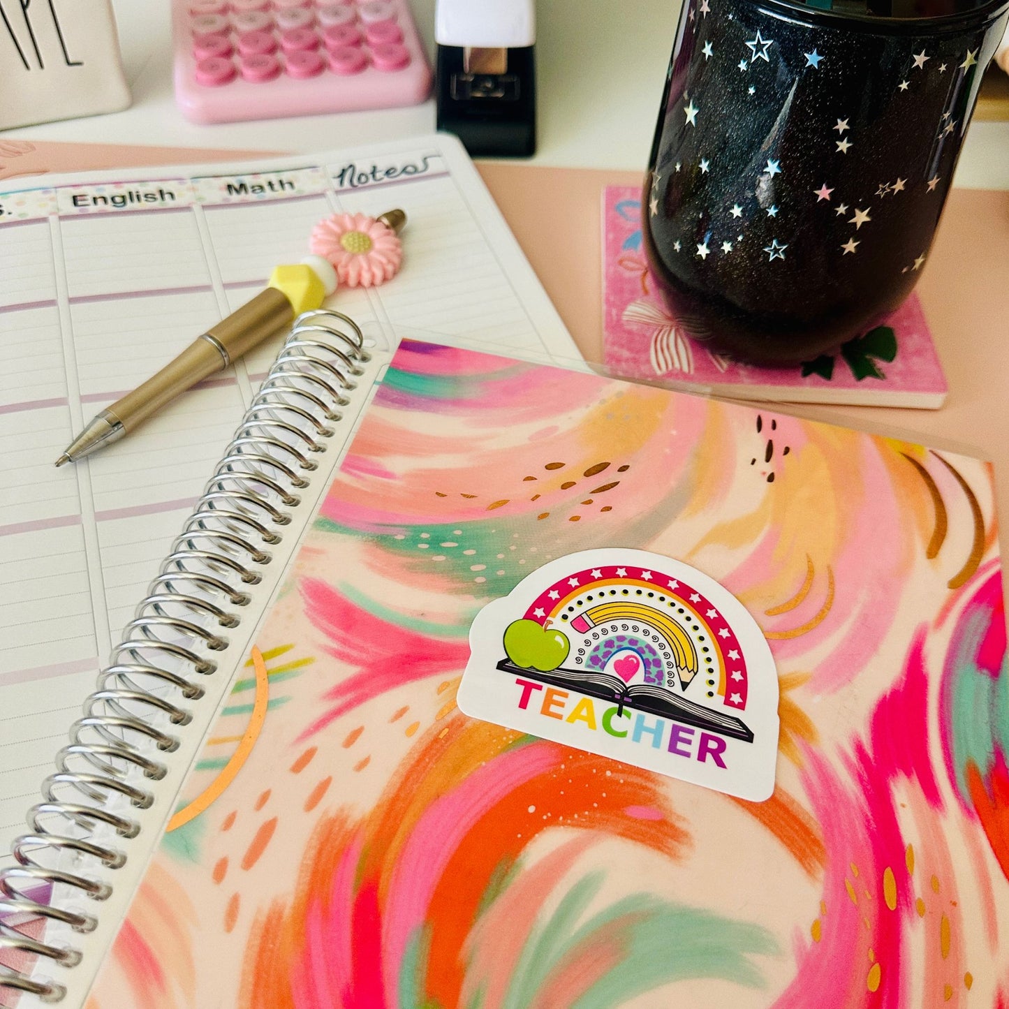 A colorful spiral-bound notebook with a pink, orange, and yellow marbled cover sits on a desk. A die-cut sticker on the cover features a rainbow, a green apple, and the word 'TEACHER'.