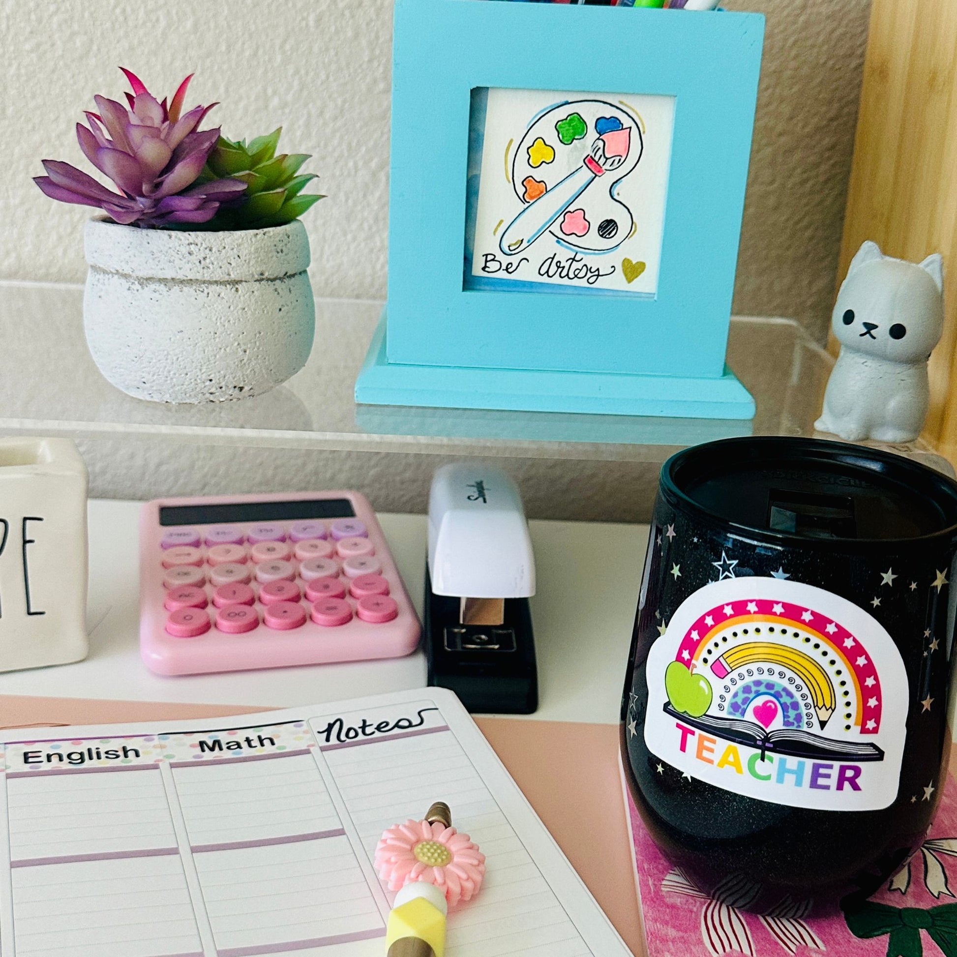 A black insulated tumbler with a colorful rainbow and 'TEACHER' sticker sits on a desk alongside a pink calculator, white stapler, planner pad, small succulent, framed art, and a white cat figurine.