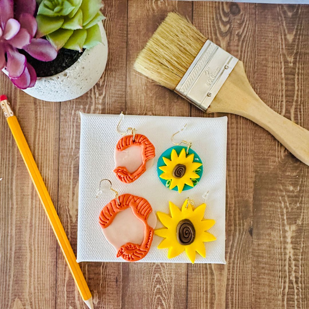 Two pairs of Van Gogh Earrings, one large set at the top and one small set at the bottom. Each pair shows an orange hair silhouette of Van Gogh and a sunflower design on a white canvas, next to a paintbrush and a succulent plant on a wooden surface.