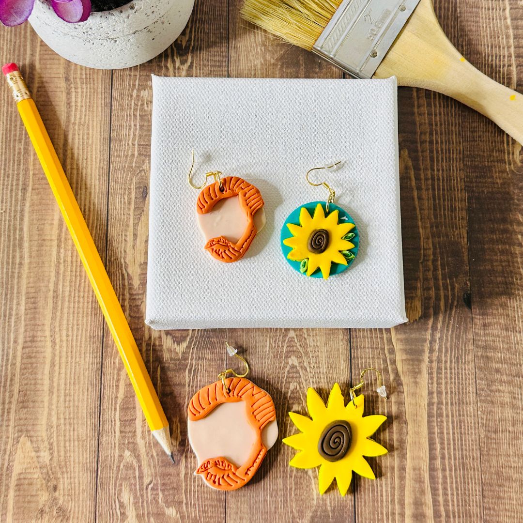 Two pairs of Van Gogh Earrings, one small set resting on top of white square canvas and below the canvas resting on the wooden table is the large Van Gogh earrings.  Each pair shows an orange hair silhouette of Van Gogh and a sunflower design on a white canvas, next to a paintbrush, a pencil, and a succulent plant on a wooden surface.