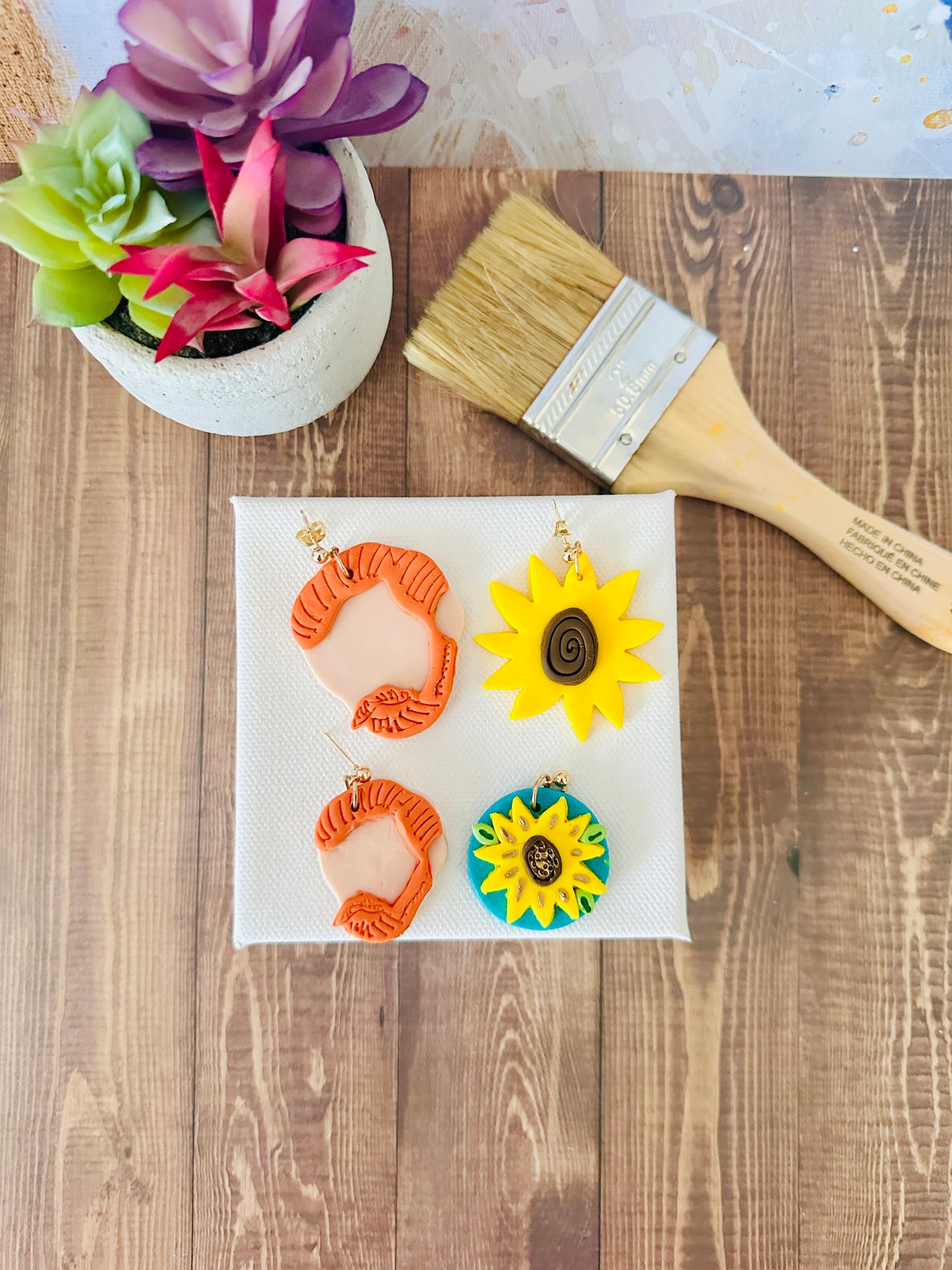 Two pairs of Van Gogh Earrings, one large set at the top and one small set at the bottom. Each pair shows an orange hair silhouette of Van Gogh and a sunflower design on a white canvas, next to a paintbrush and a succulent plant on a wooden surface.