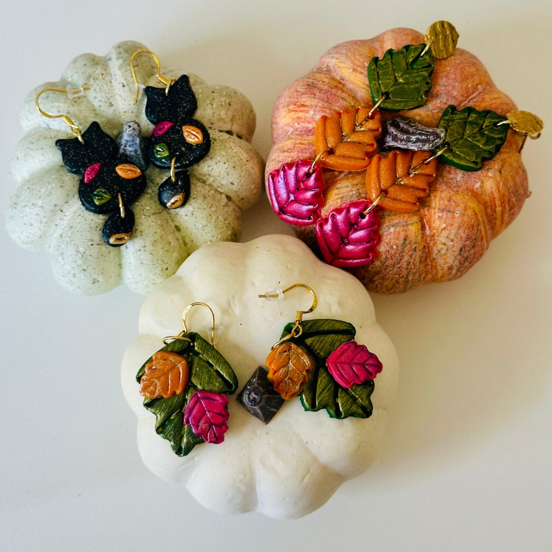 Three pairs of handmade autumn-themed earrings displayed on decorative pumpkins against a plain white background. The earrings feature colorful polymer clay leaves from cranberry, pumpkin orange, and forest green to a black cat with colorful fall leaves. Each pair has gold hoops and another with bronze earring clasp.