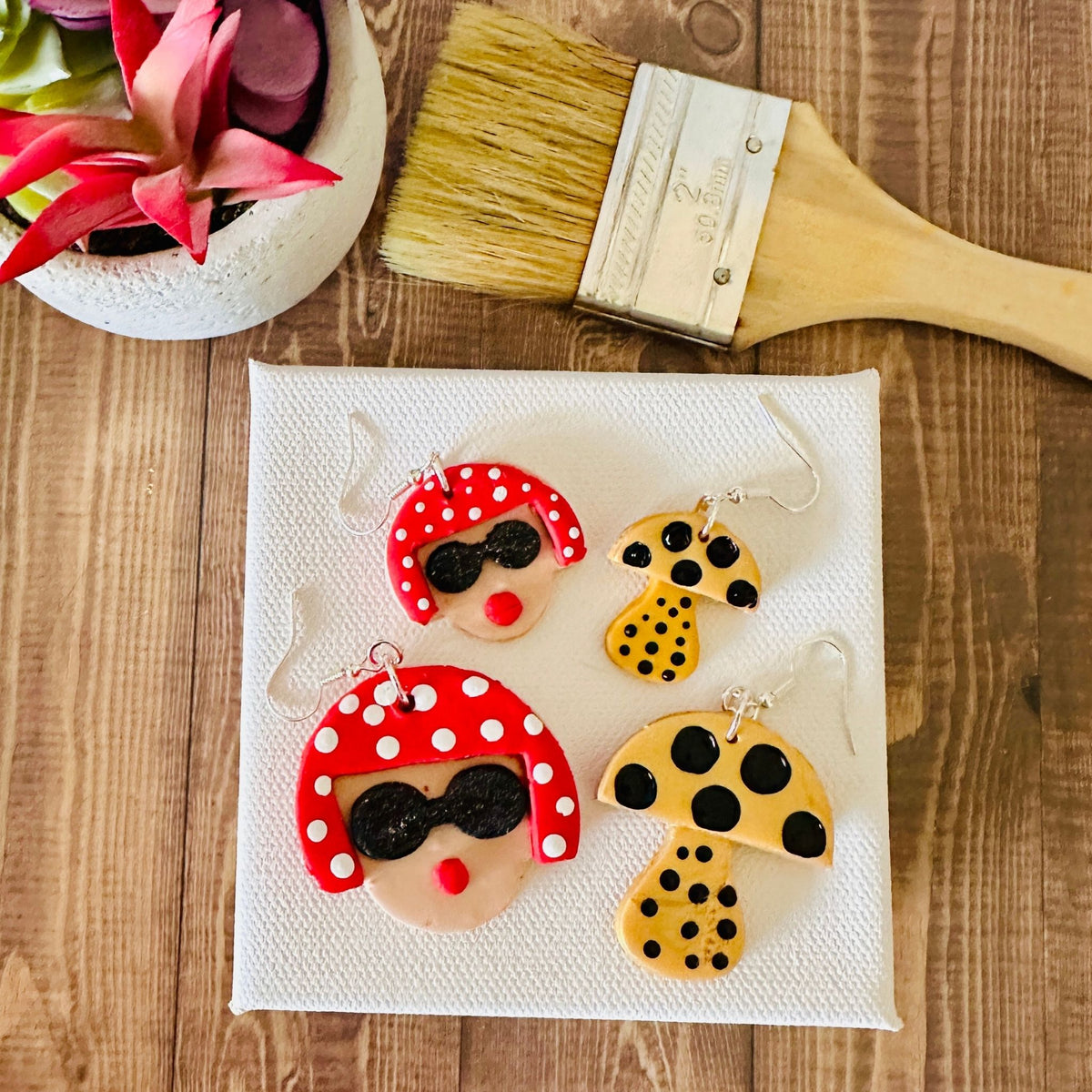 Large and small Yayoi Kusama earrings with portrait and mushroom design on white canvas with paintbrush and potted plant on wooden table.