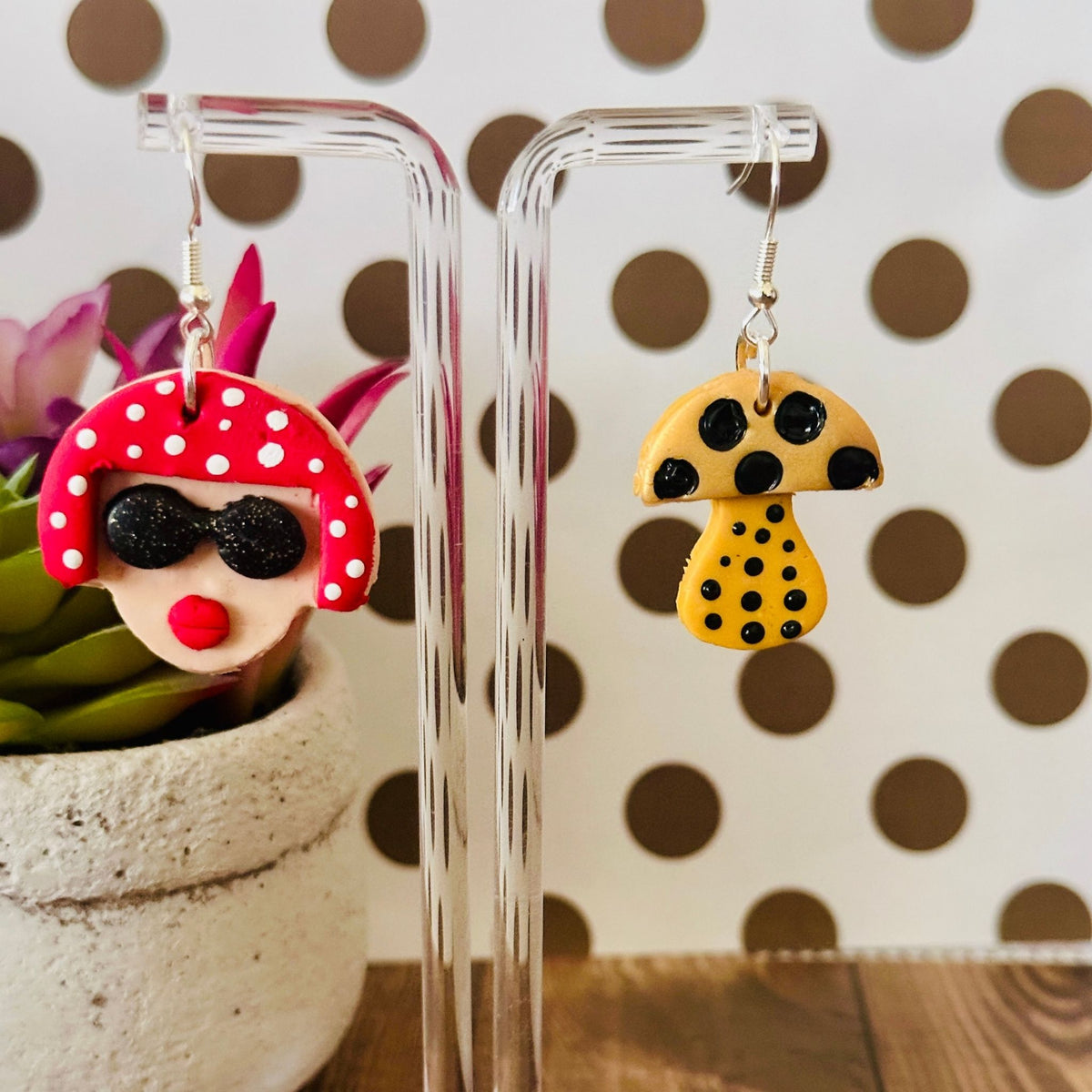 Close-up of Yayoi Kusama polymer clay earrings on clear stand with polka dot background and wooden table, highlighting handcrafted details.