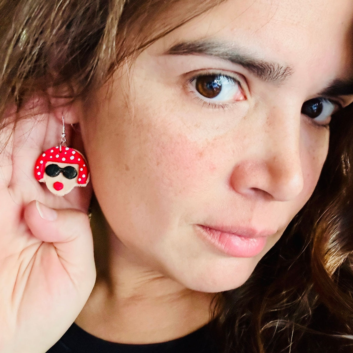Woman wearing Yayoi Kusama polymer clay earrings displaying the portrait earring with red polka dot hair, black sunglasses and red lips.
