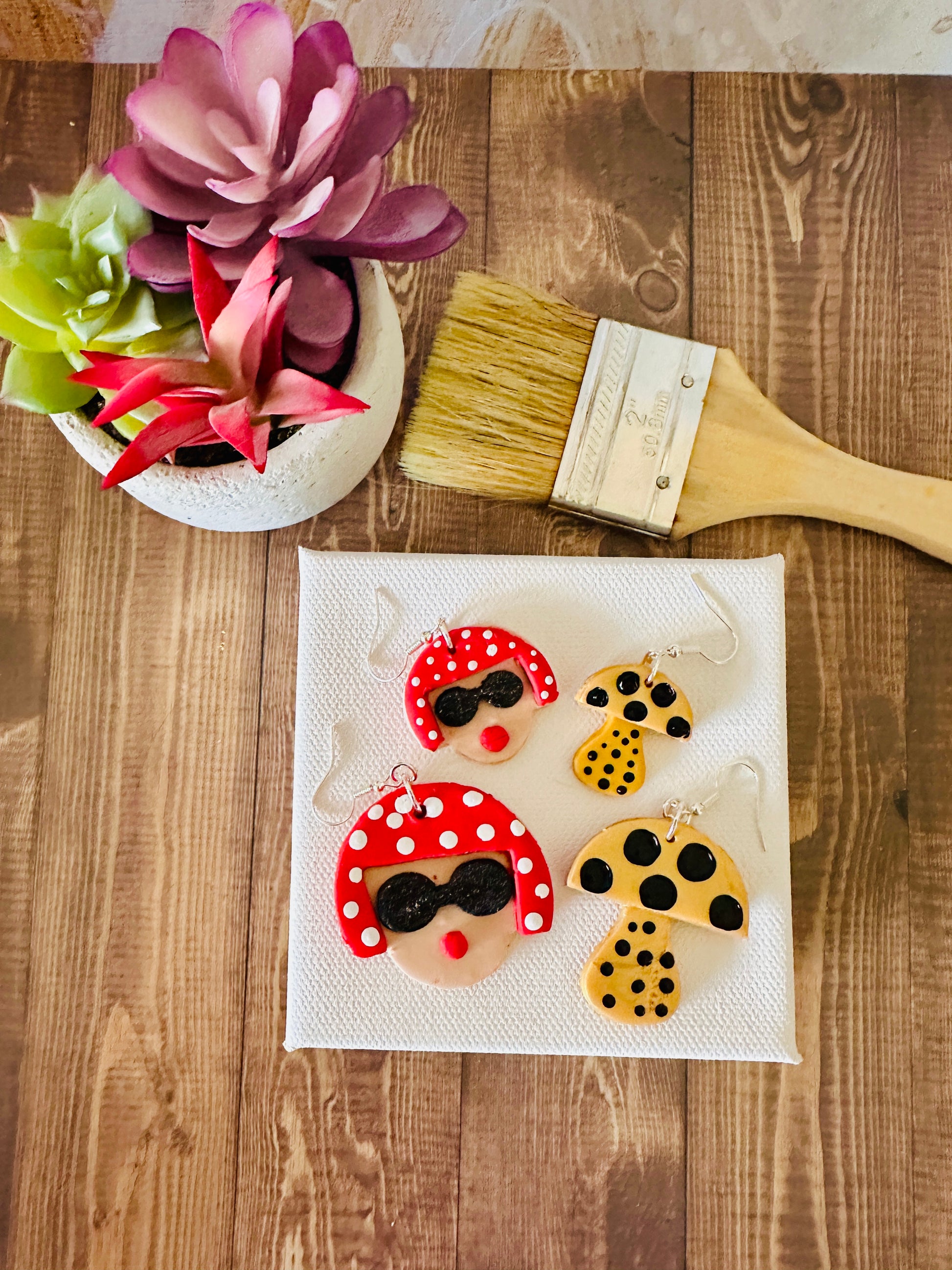 Large and small Yayoi Kusama earrings with portrait and mushroom design on white canvas with paintbrush and potted plant on wooden table.