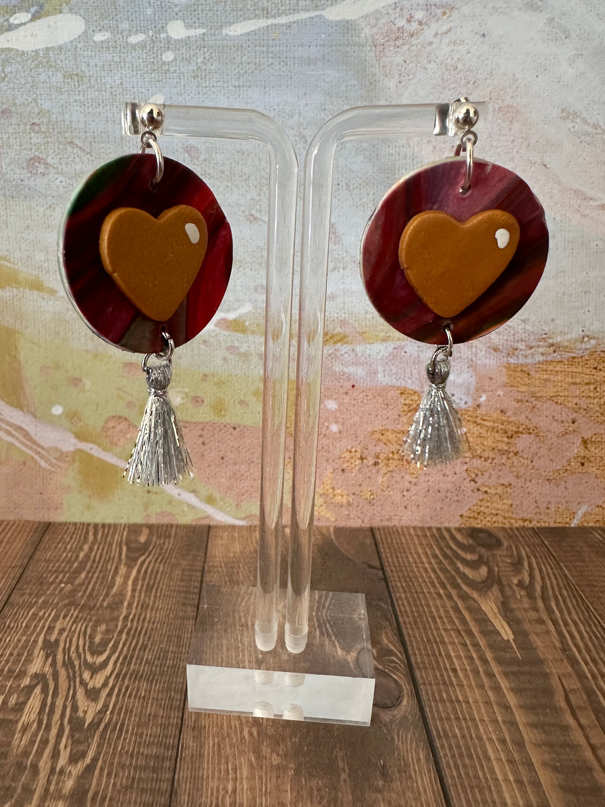Handmade polymer clay heart earrings on a clear earring stand in front of a colorful abstract wall.
