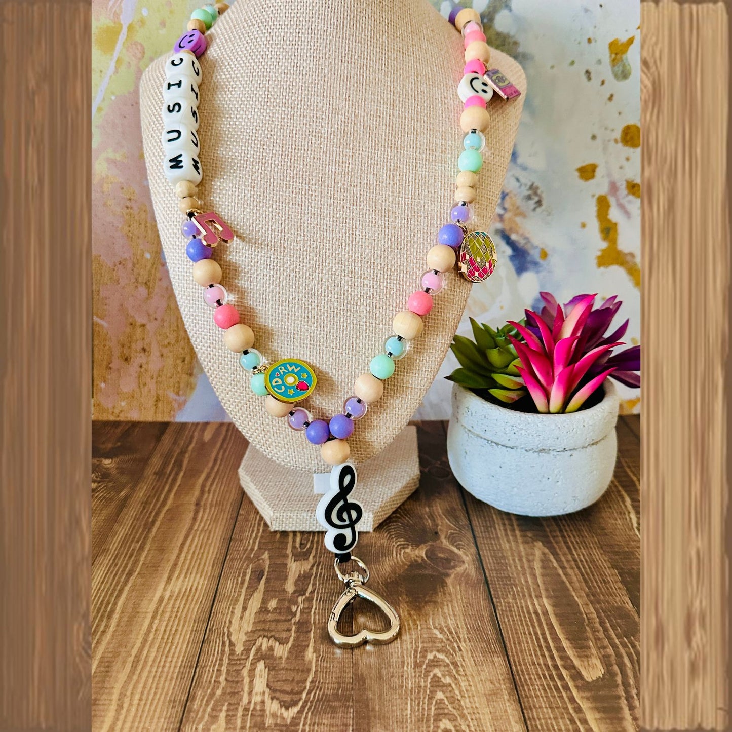 Colorful beaded necklace with music-themed charms displayed on a wooden base, next to a small potted plant.