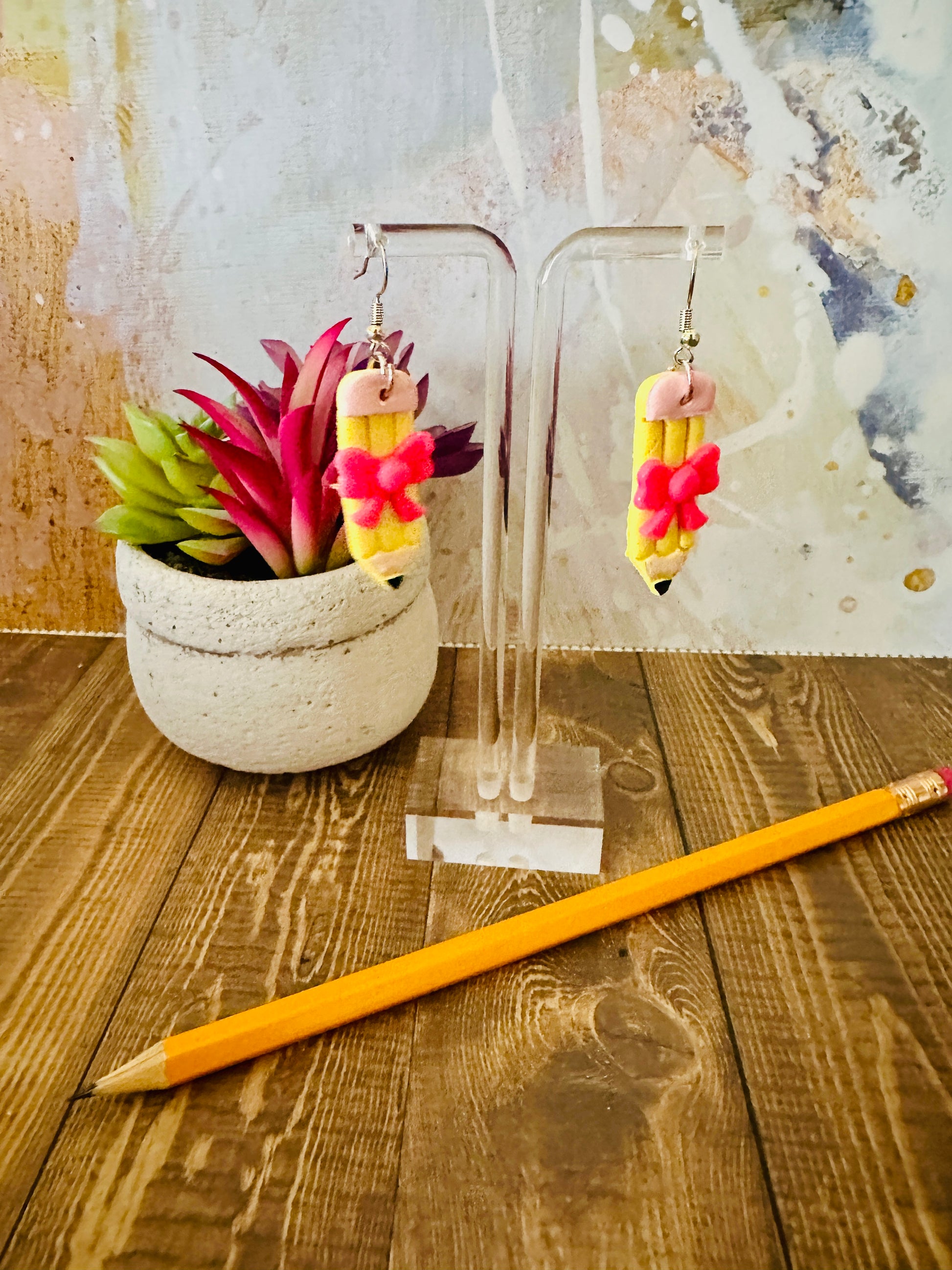 Straight pencil earring on stand next to a real pencil for size reference, wooden table and colorful wall background.