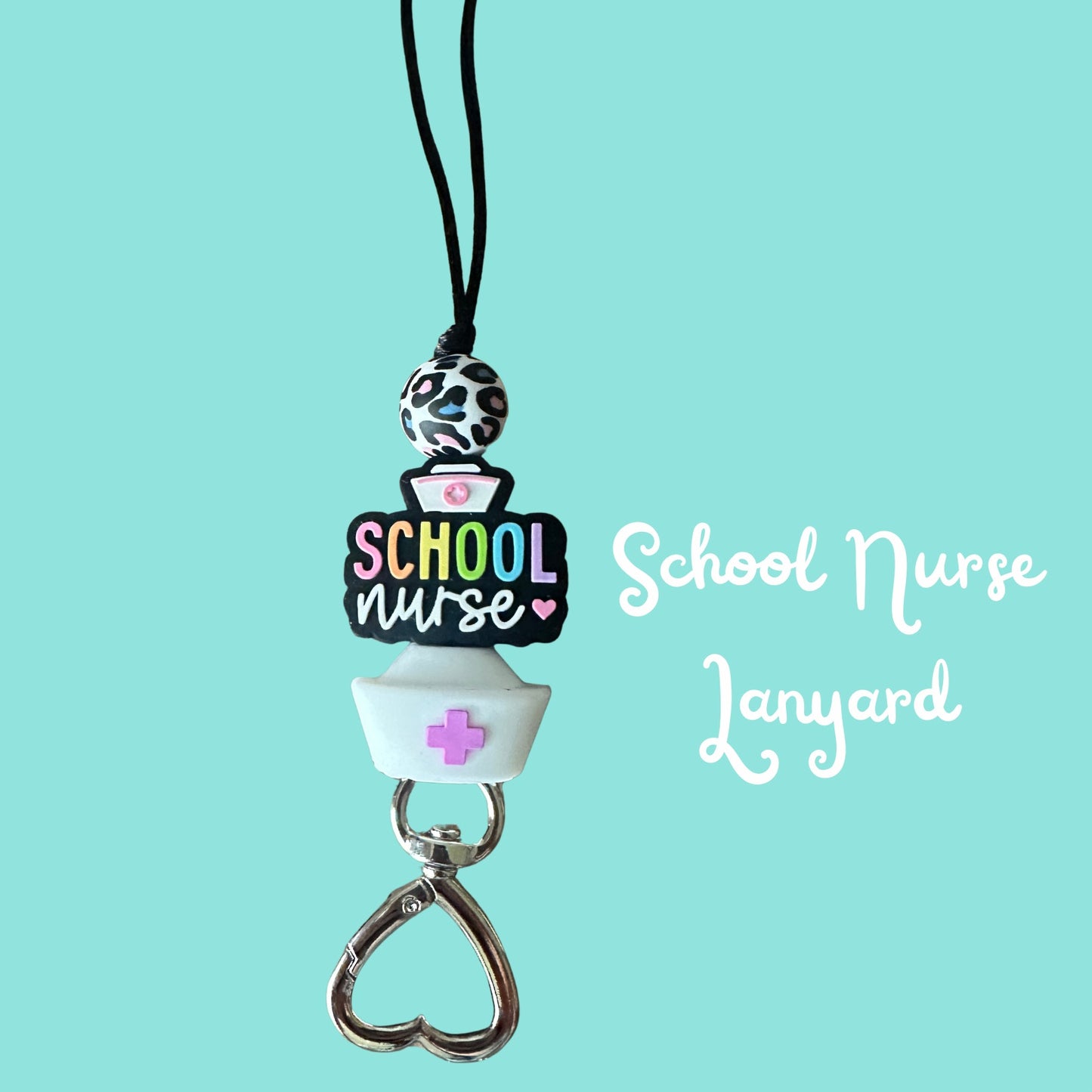 A school nurse themed or lanyard on a black cord, featuring a black and white patterned top bead, a colorful 'SCHOOL NURSE' sign, a white nurse hat, and a silver heart-shaped clasp, set against a solid light blue background with white text reading 'School Nurse Lanyard'.
