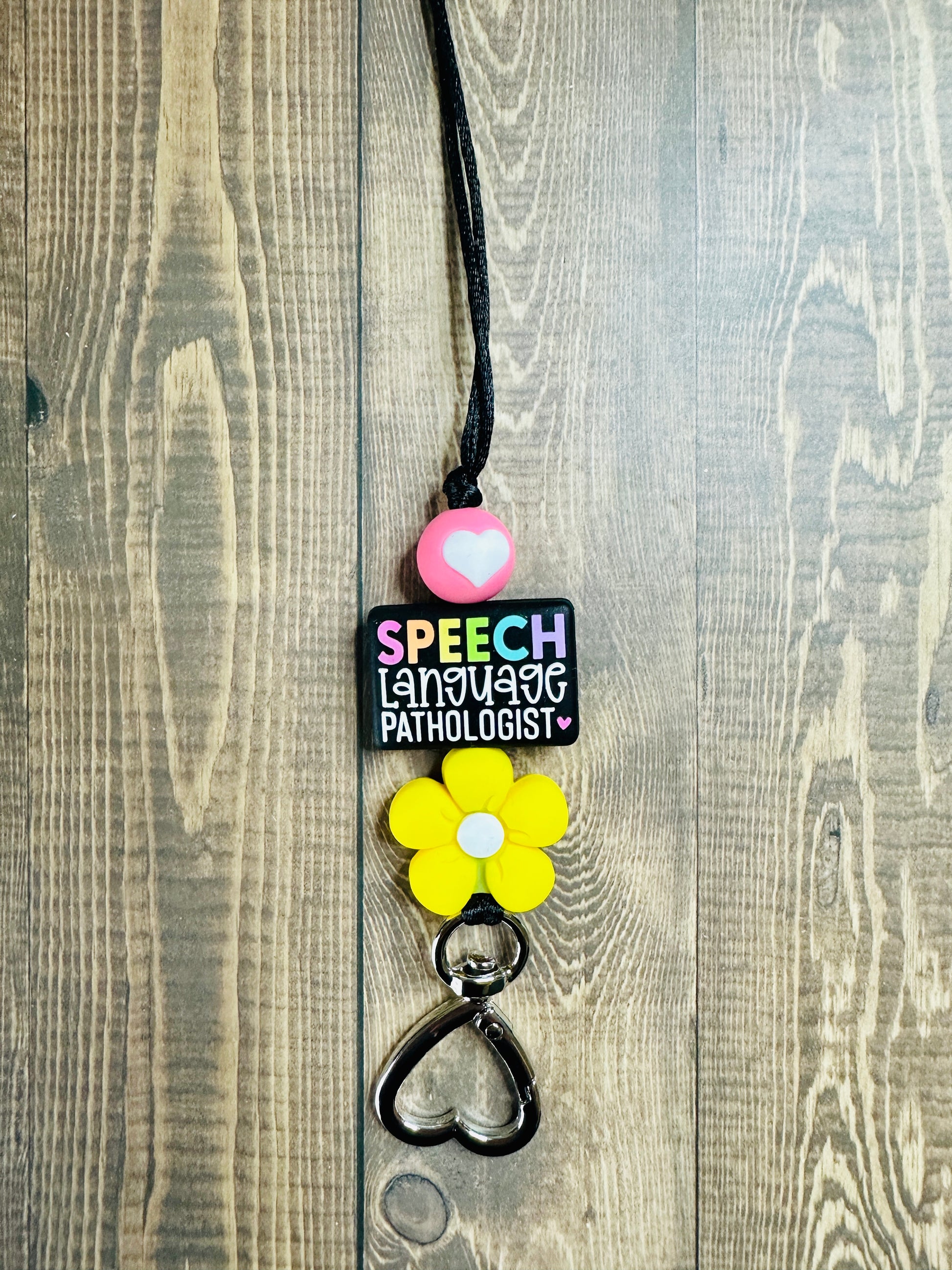 Detail view of handmade speech language pathologist lanyard on wooden table featuring rainbow SPEECH silicone focal bead and heart-shaped clasp.